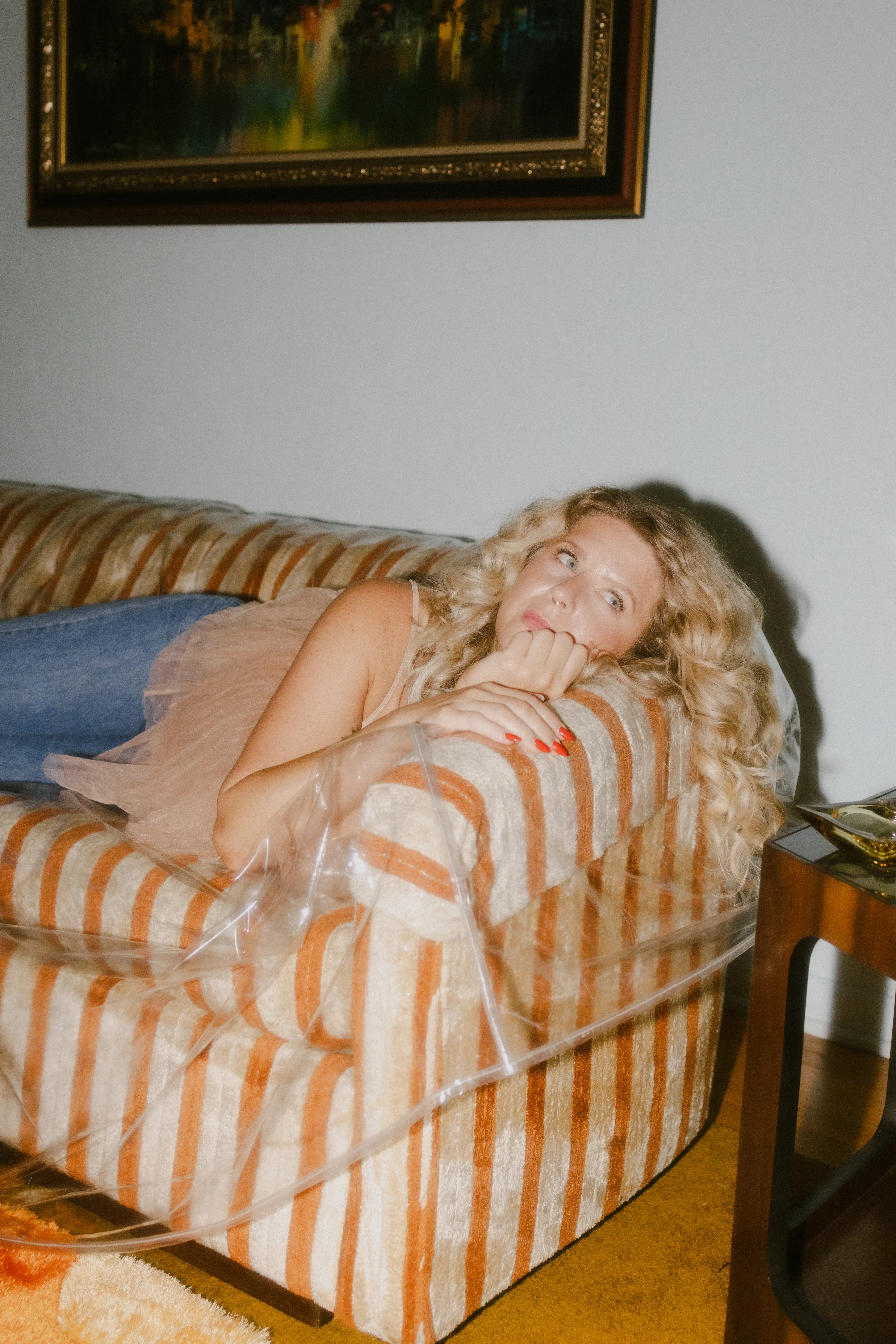 Kathleen Whittemore lying on a striped orange and beige couch covered with clear plastic, resting her chin on her hand.