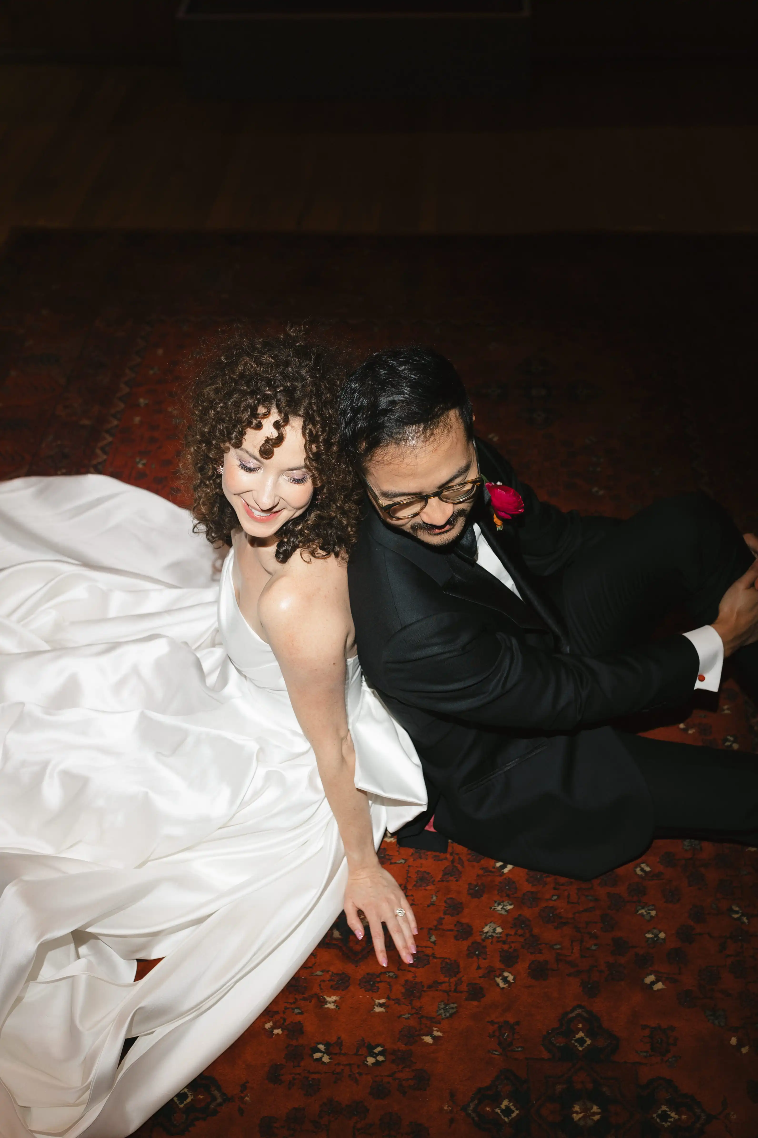 Bride in a white wedding dress and groom in a black suit sitting back-to-back on a red patterned rug, smiling and looking down.