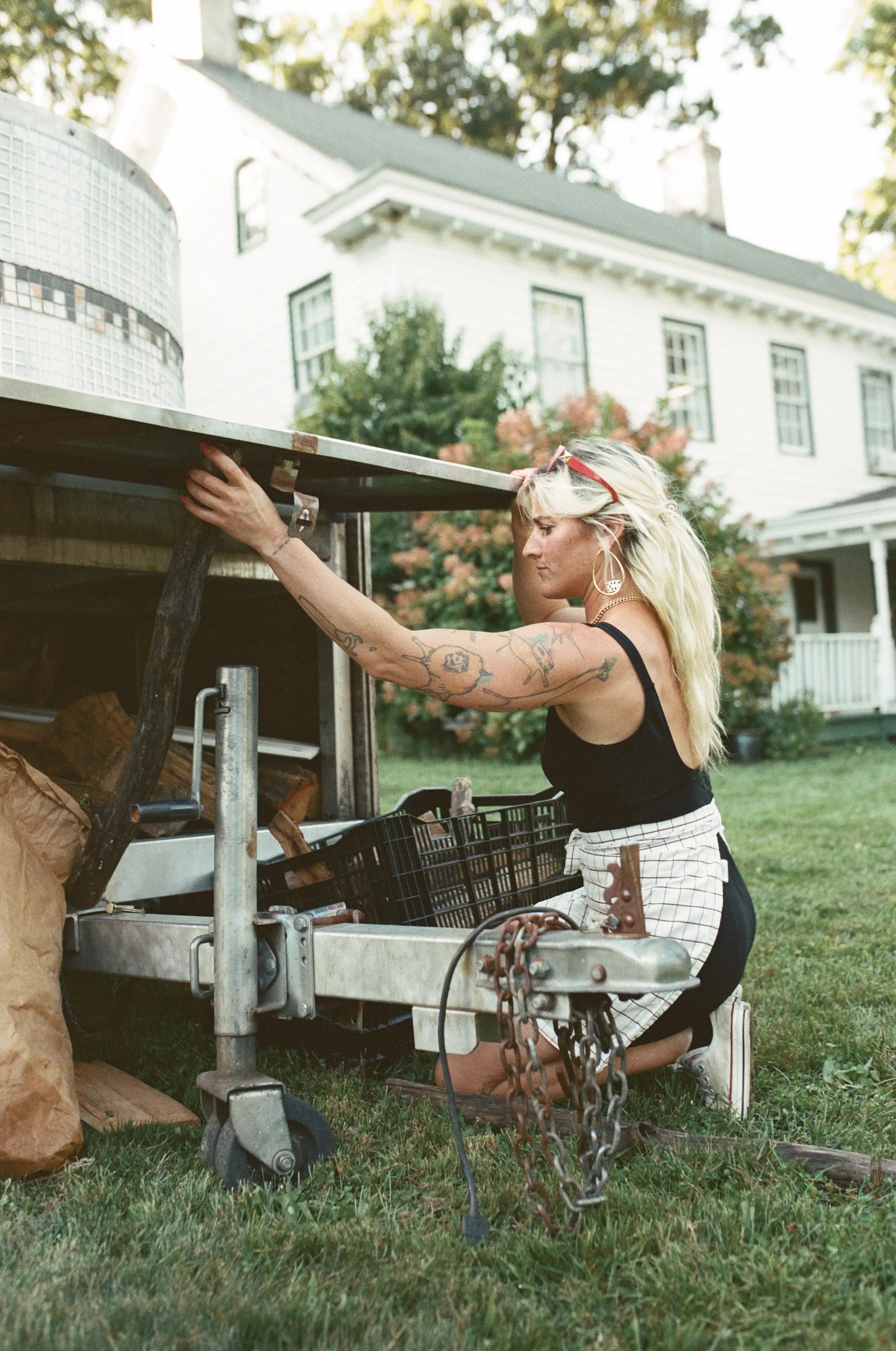 Tattooed woman with blonde hair kneeling on grass, opening a metal trailer side next to a house.