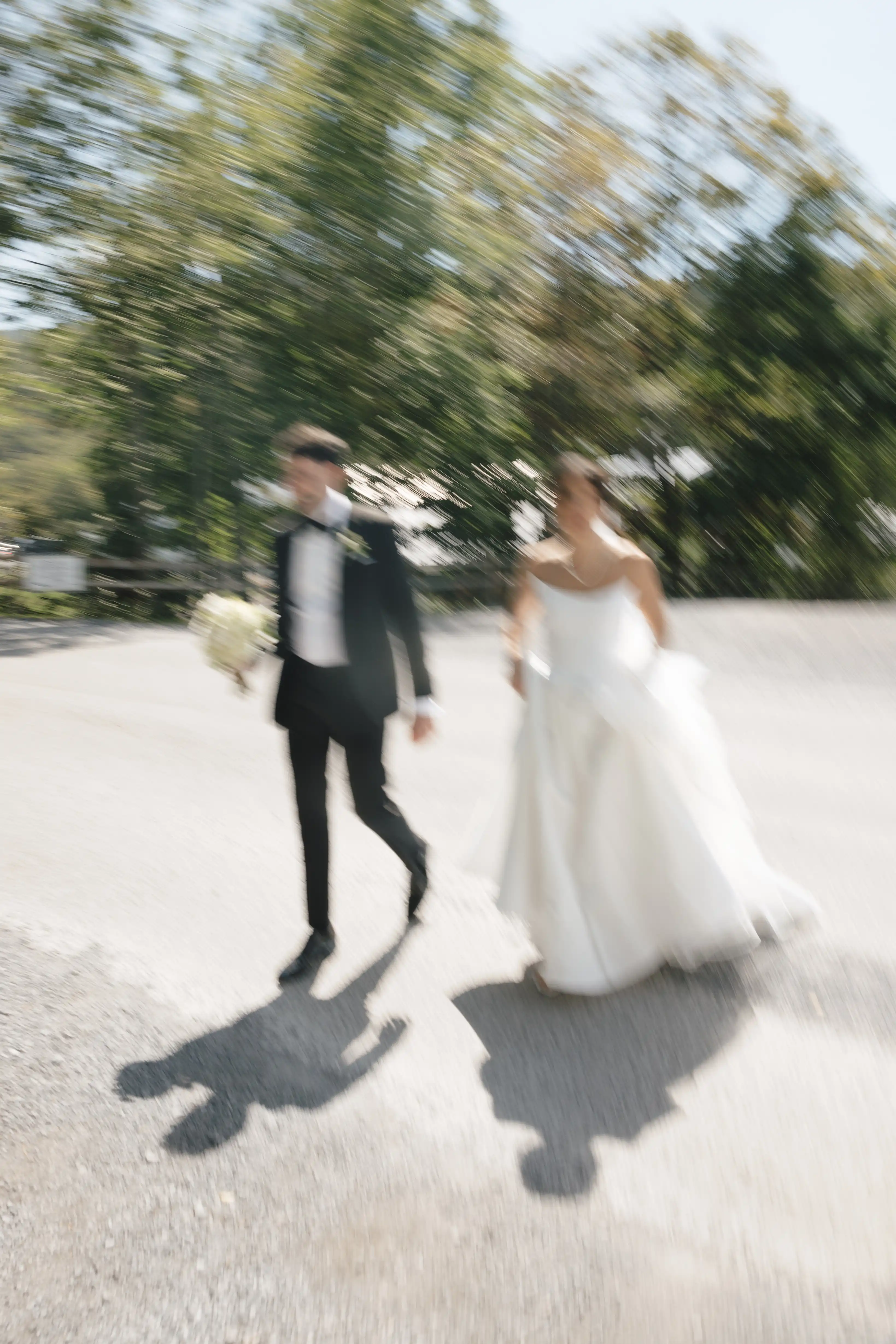 Blurred image of a bride in a white dress and a groom in a black suit walking outdoors on a sunny day.