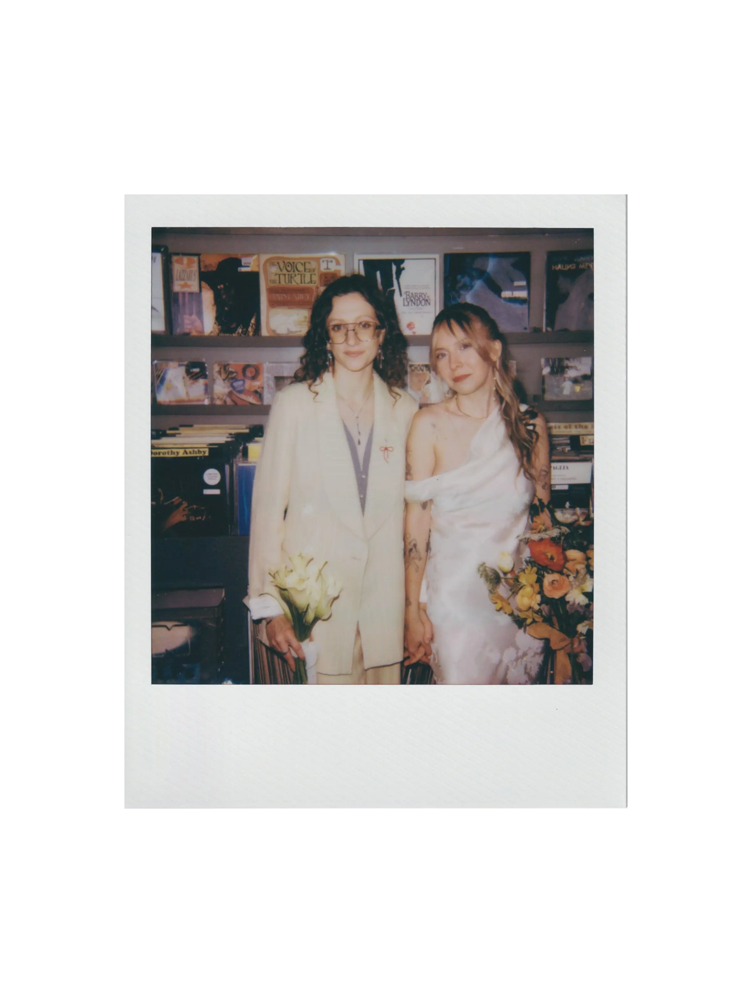 Polaroid photo of two women holding hands, one in a cream suit holding white calla lilies, the other in a white one-shoulder dress holding a colorful bouquet, standing inside a record store.