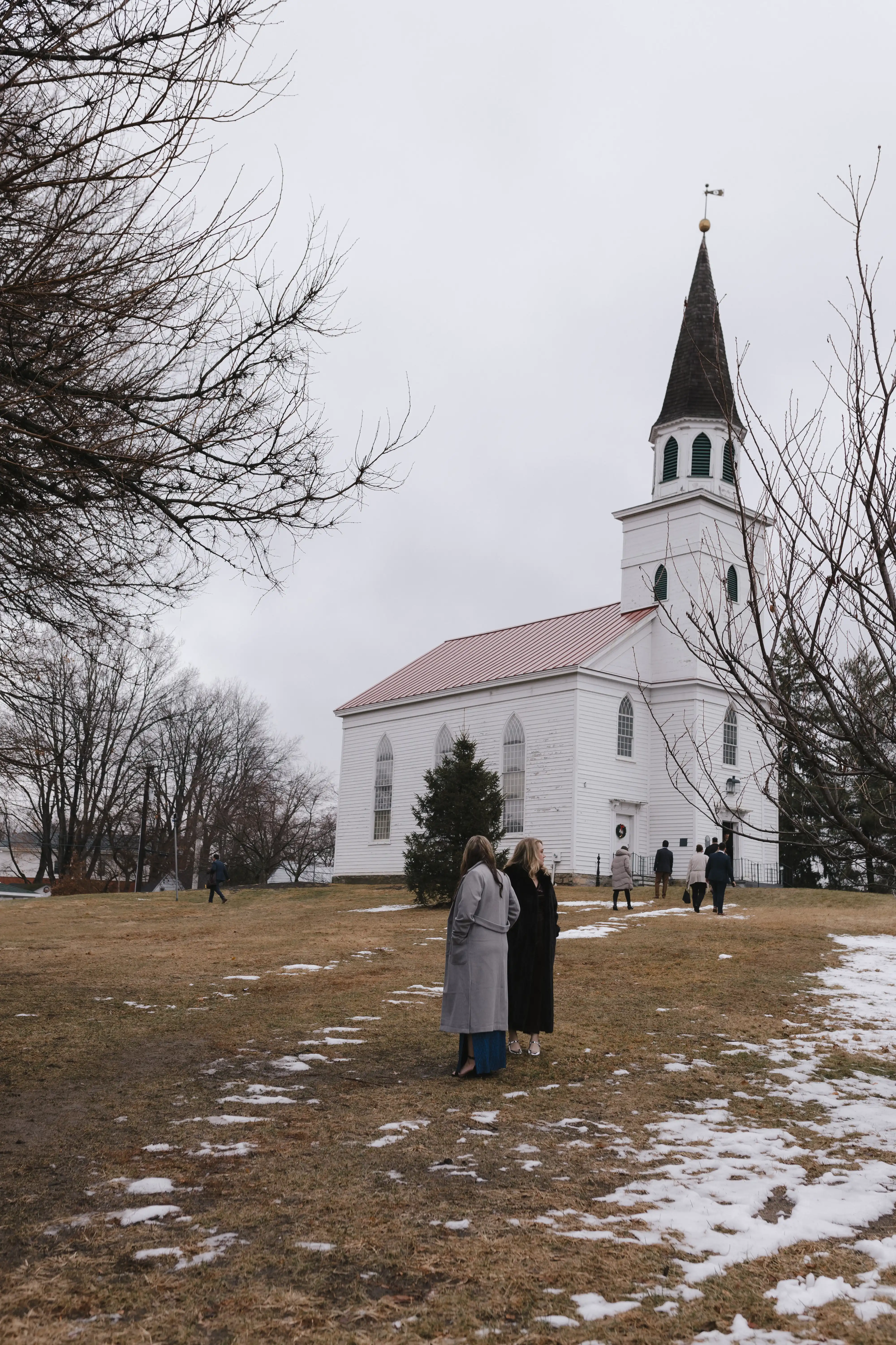 People in coats standing and walking near a white church with a tall steeple on a grassy area with patches of snow under an overcast sky.