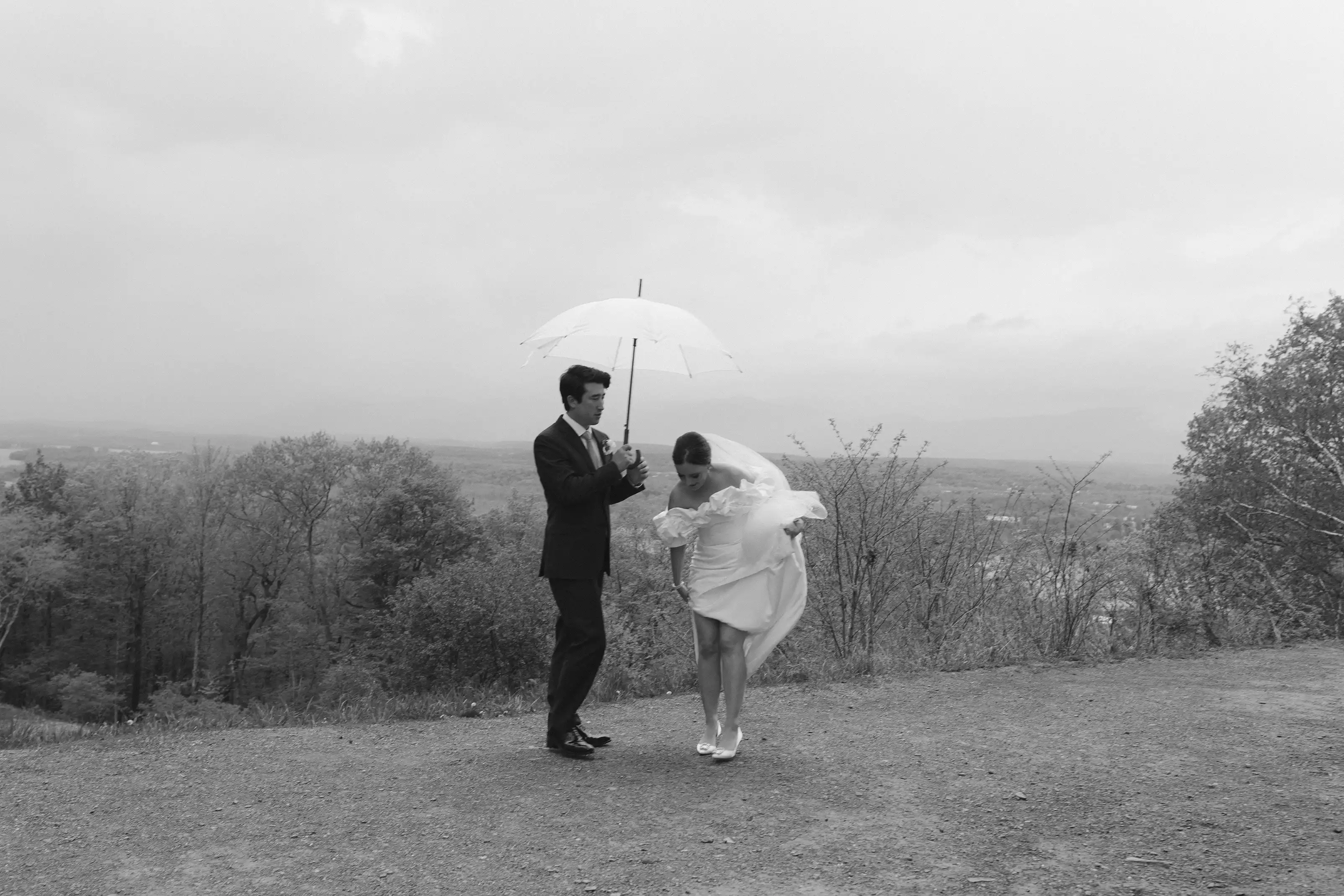 Groom holding an umbrella over bride outdoors on a cloudy day with trees and landscape in the background.