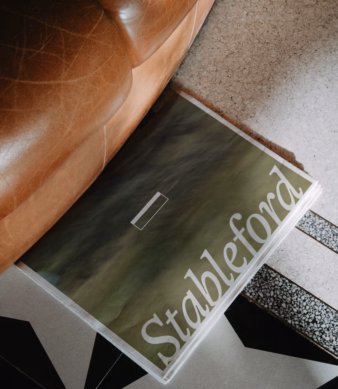 Newspaper titled 'Stableford' partially tucked under a brown leather couch on a tiled floor.