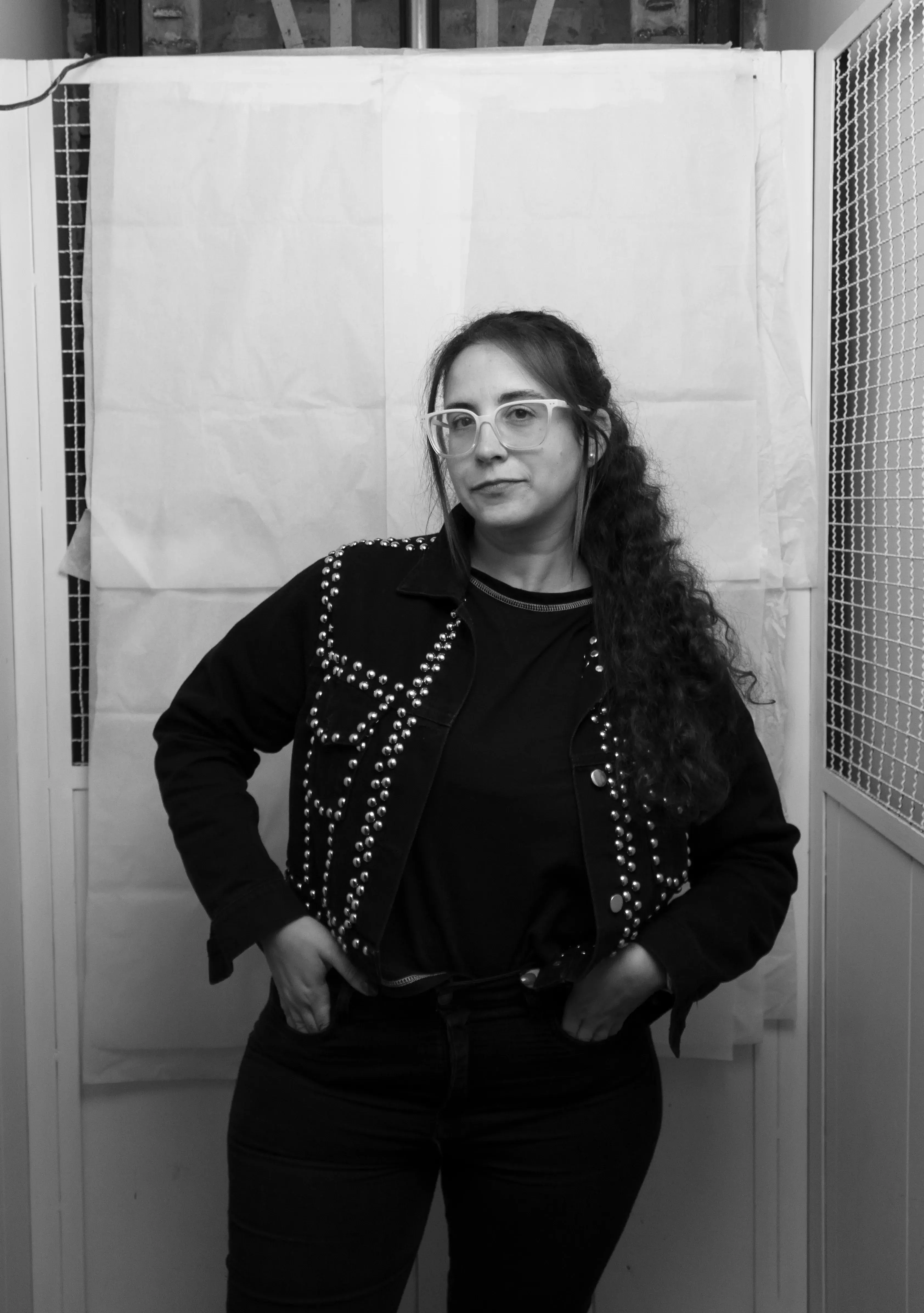 Portrait of a woman wearing glasses and a studded jacket standing with hands in pockets against a simple backdrop.