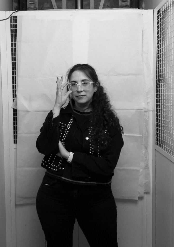 Woman with long curly hair adjusting white-framed glasses, wearing a studded jacket and standing in front of a plain backdrop.