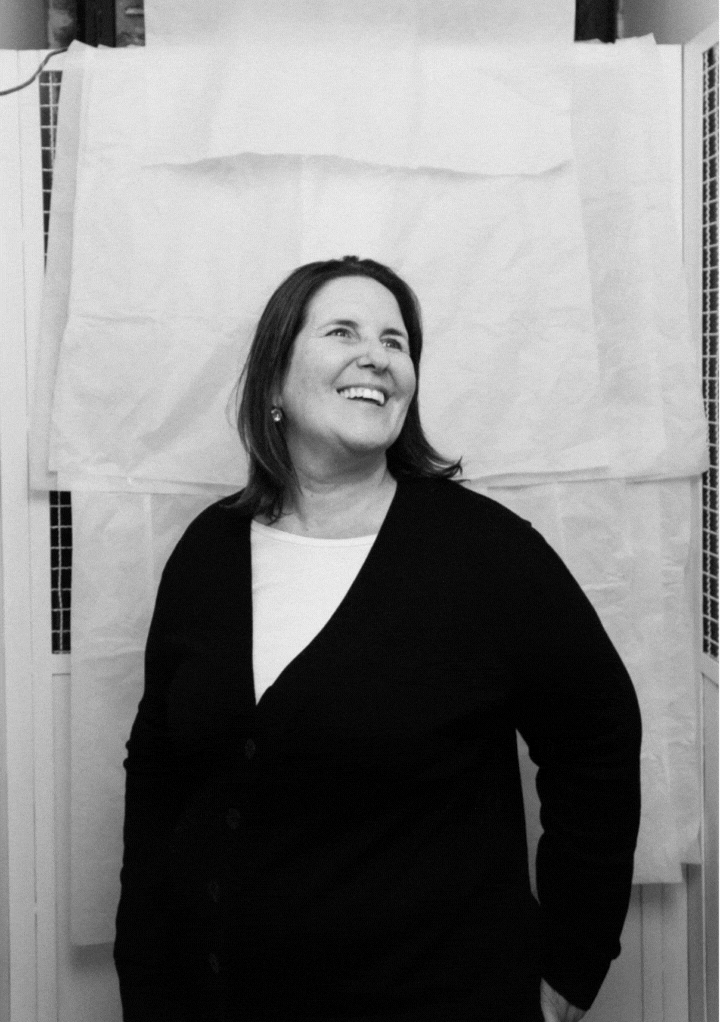 Smiling woman in a black cardigan standing against a backdrop of white sheets.