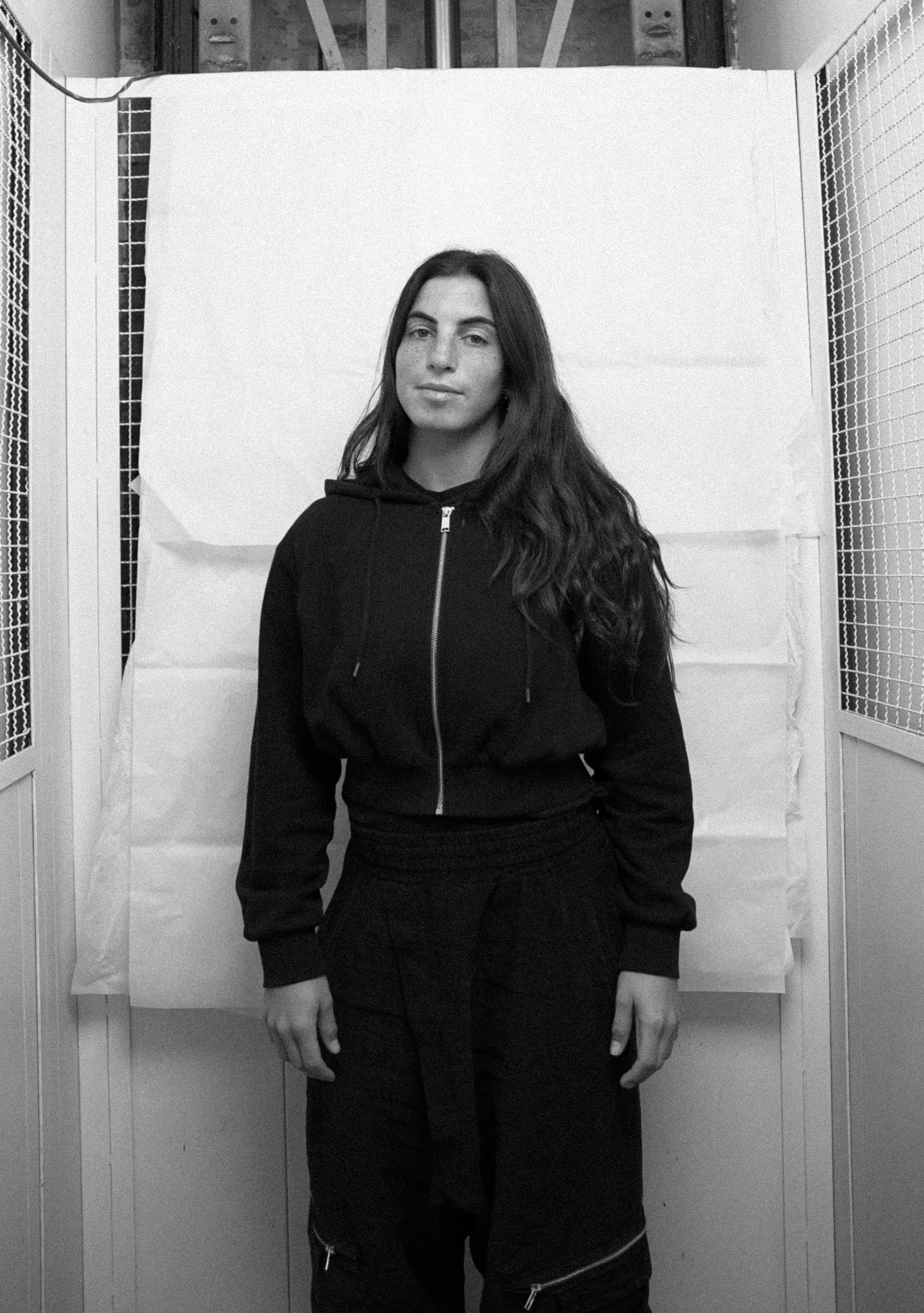 Woman with long dark hair wearing a black zip-up hoodie and pants standing against a white backdrop in a narrow space with mesh panels.