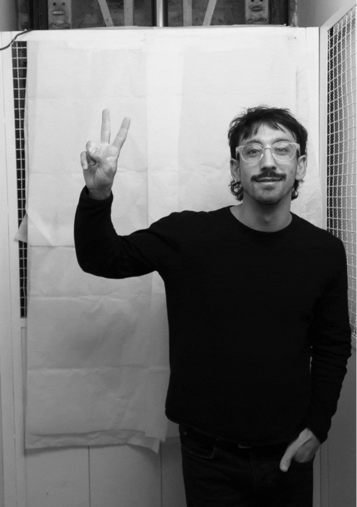 Man with glasses and a mustache wearing a dark sweater making a peace sign with his right hand.