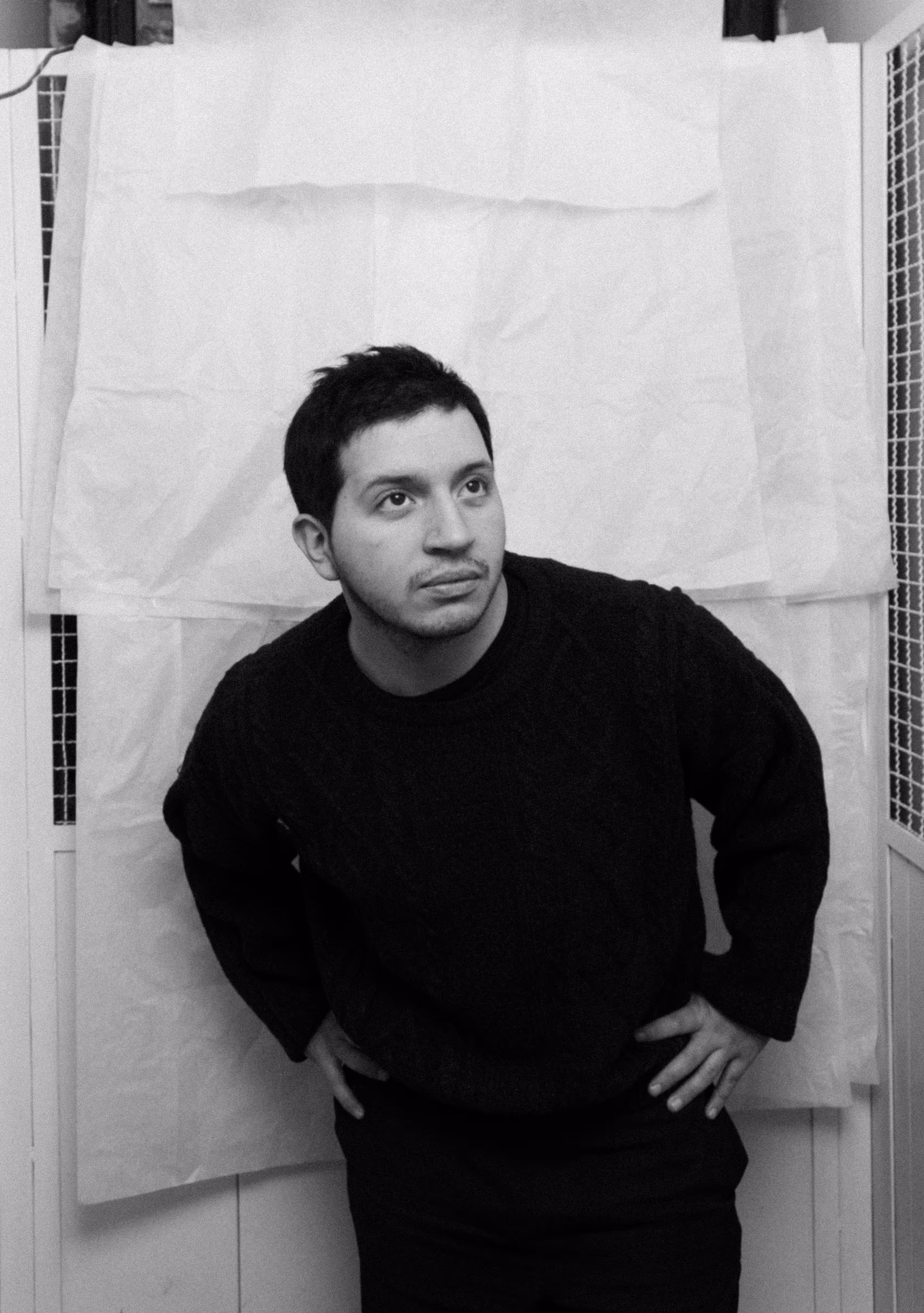 Man with short dark hair wearing a dark sweater leans forward with hands on hips against a white background.