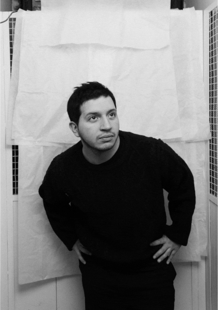 Black and white photo of a man with short dark hair and a beard, wearing a dark sweater, standing with hands on hips in front of a plain draped background.