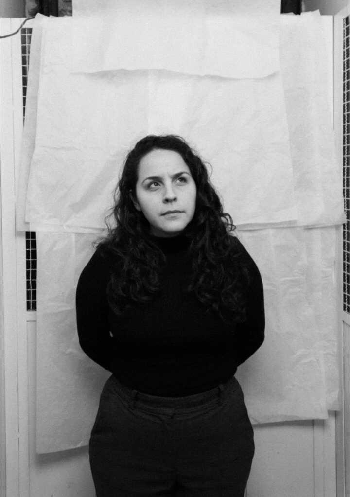 Black and white photo of a man in a dark sweater standing with hands on hips, looking slightly upward and to the side.