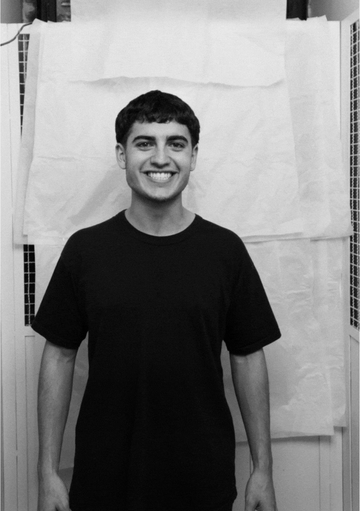 Smiling young man with short dark hair wearing a plain black t-shirt standing in front of a light-colored background.