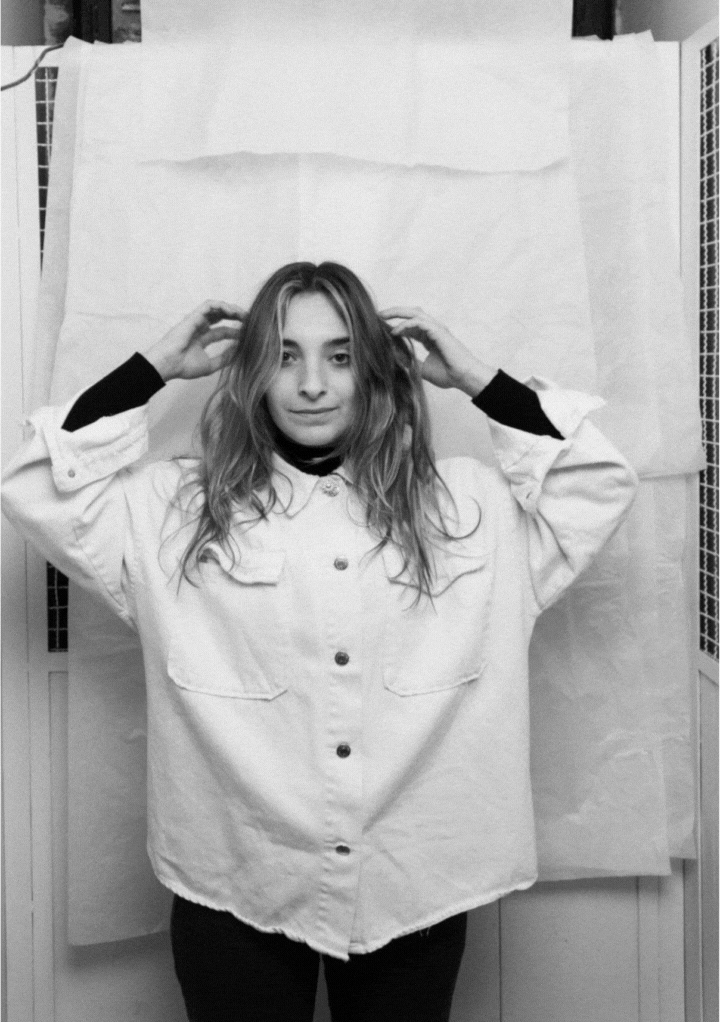 Black and white photo of a woman with long hair wearing a loose button-up shirt, holding her hair with both hands.