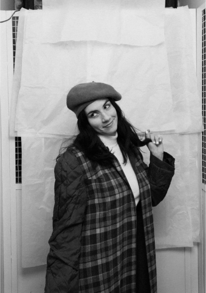 Black and white photo of a woman wearing a beret and a plaid coat, playfully holding a lock of her hair and looking to the side.