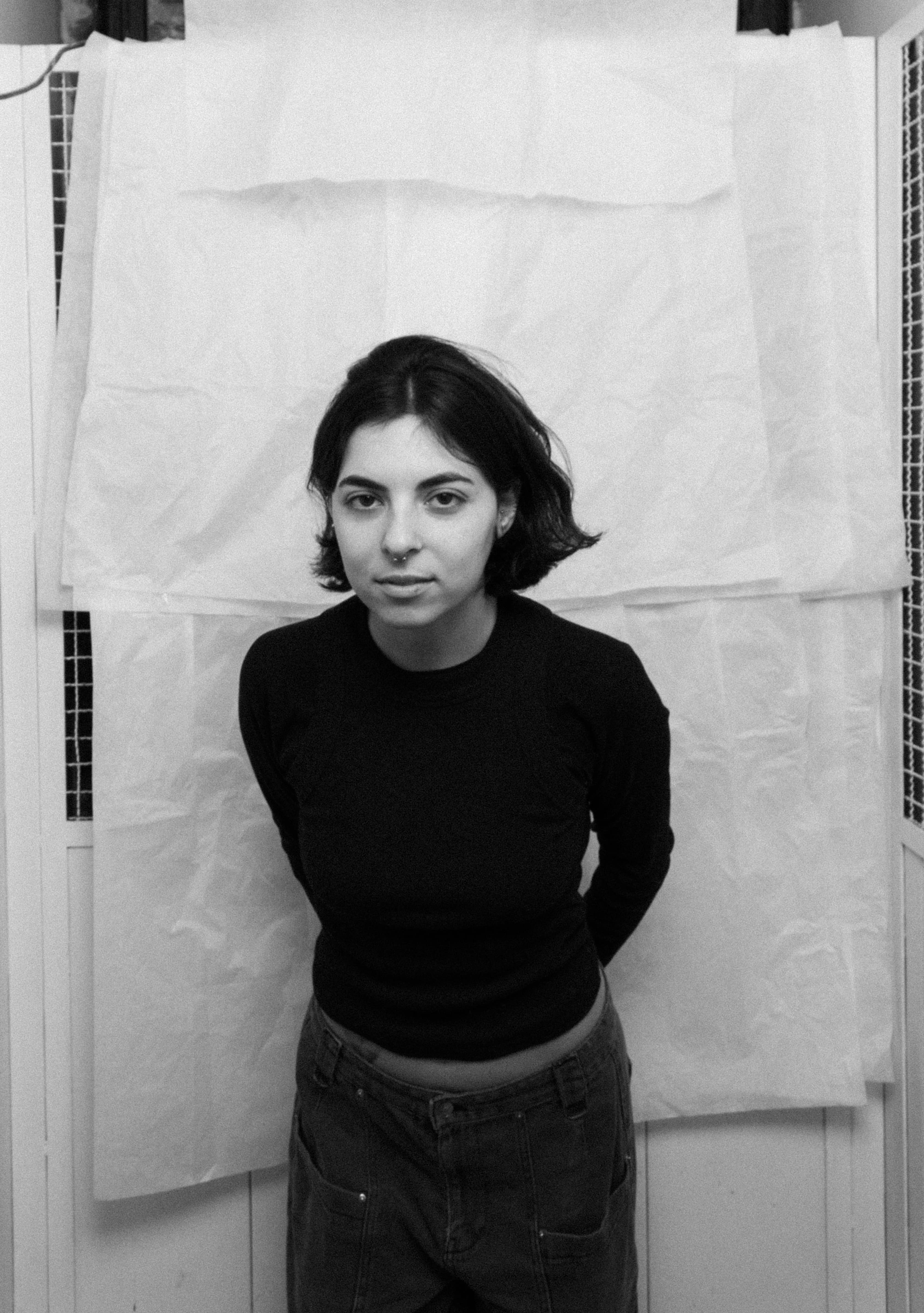 Black and white portrait of a woman with short hair wearing a black long-sleeve shirt and dark pants standing with hands behind her back.