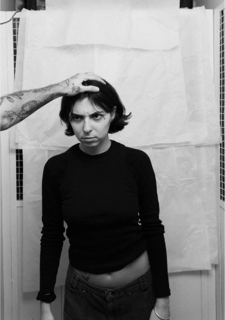 Black-and-white photo of a serious young woman with short hair wearing a black long-sleeve shirt, a tattooed arm touching her head.