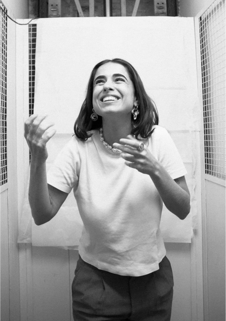 Black and white photo of a smiling woman with short hair, wearing a white t-shirt, pearl necklace, and earrings, gesturing with her hands.