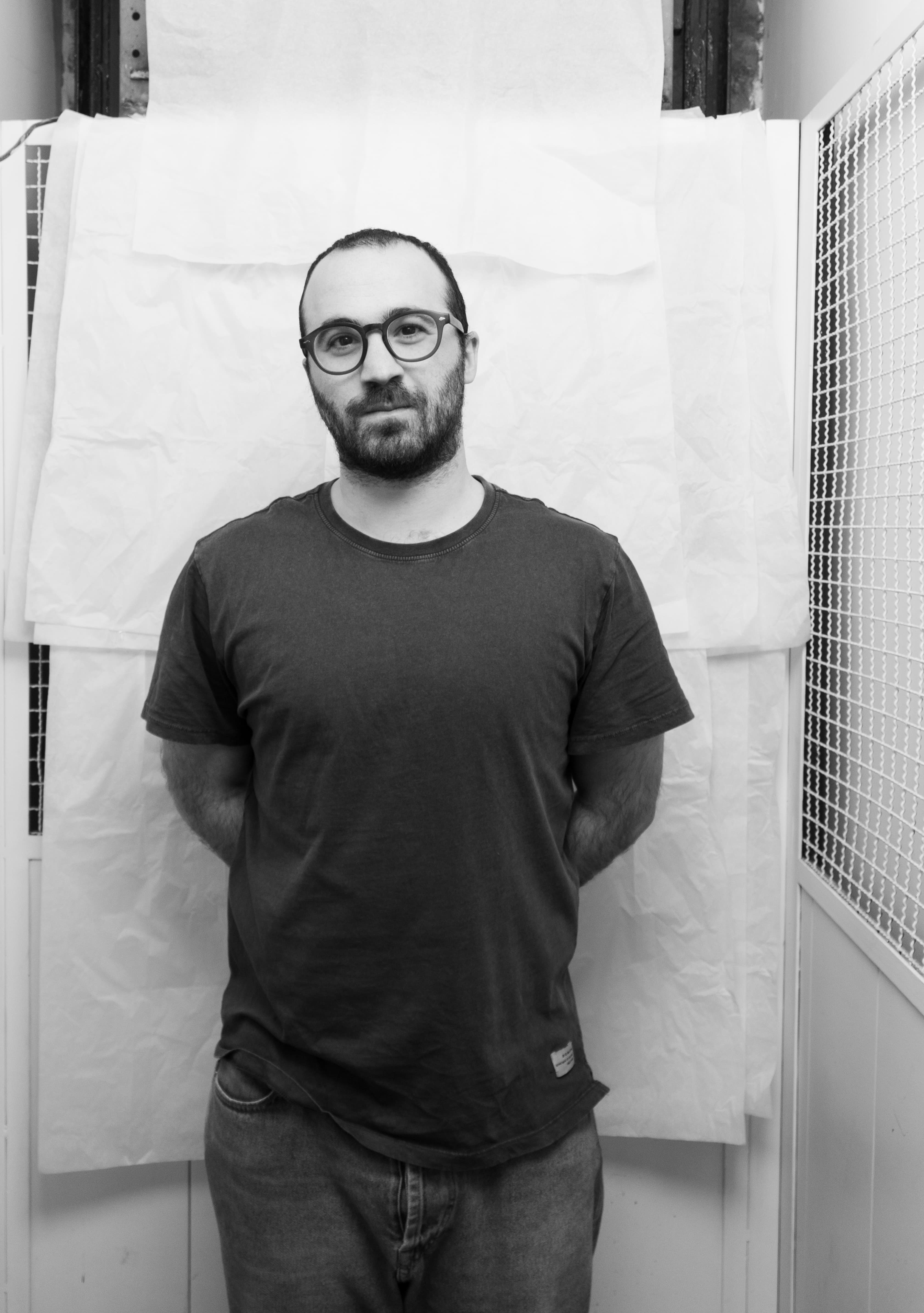 Portrait of a man with glasses and a beard standing with hands behind his back in front of a white textured backdrop.