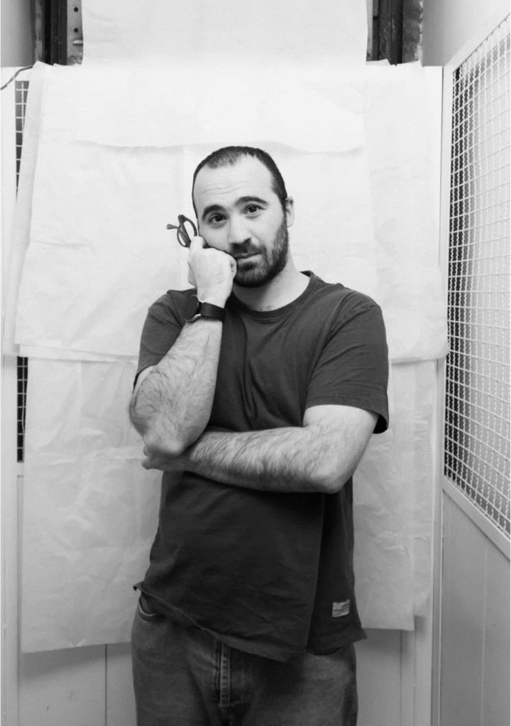 Black and white photo of a man with a beard holding glasses to his face, standing with crossed arms in front of a textured backdrop.
