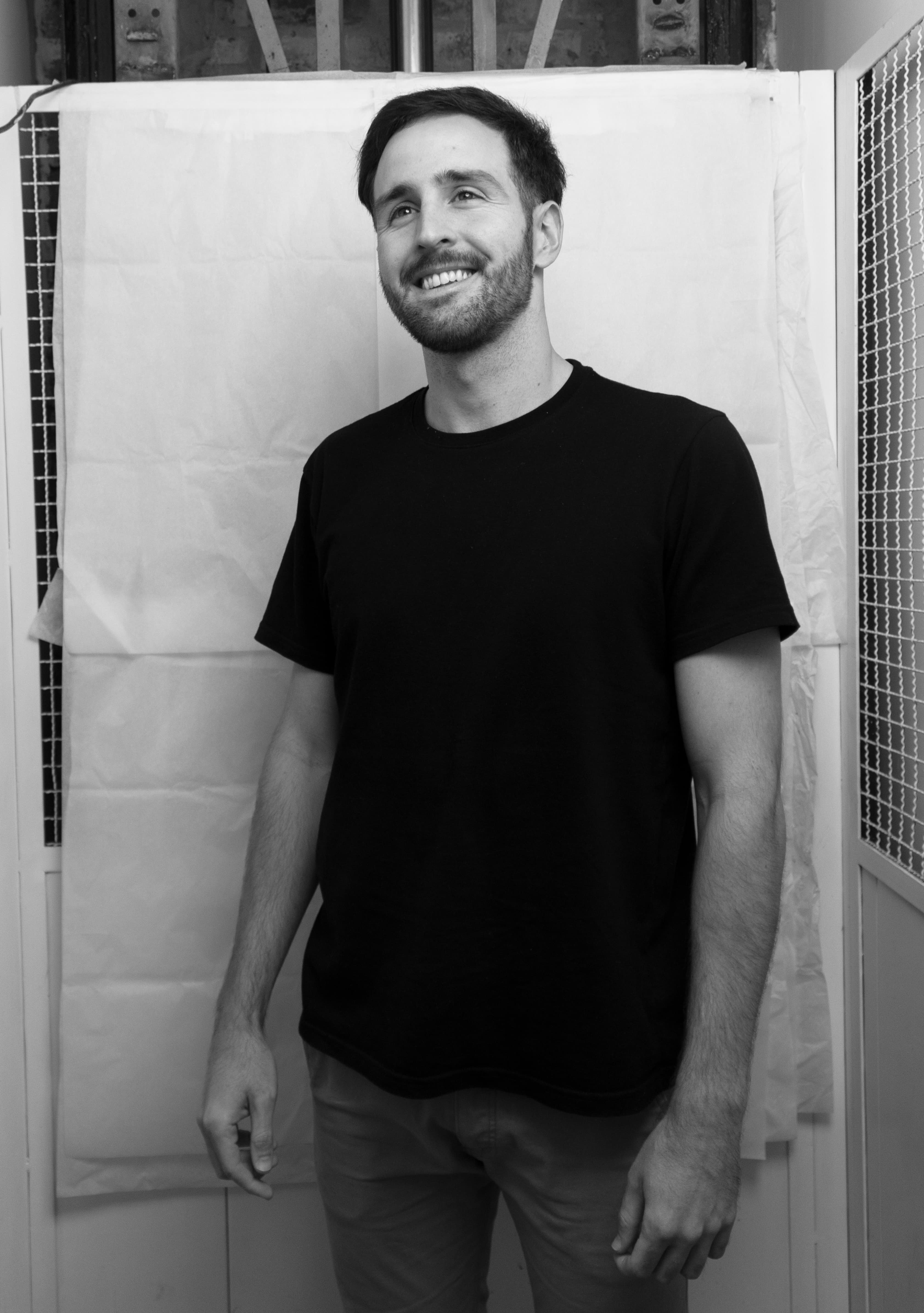 Smiling man with short dark hair and beard wearing a black t-shirt standing against a white paper backdrop in a black and white photo.