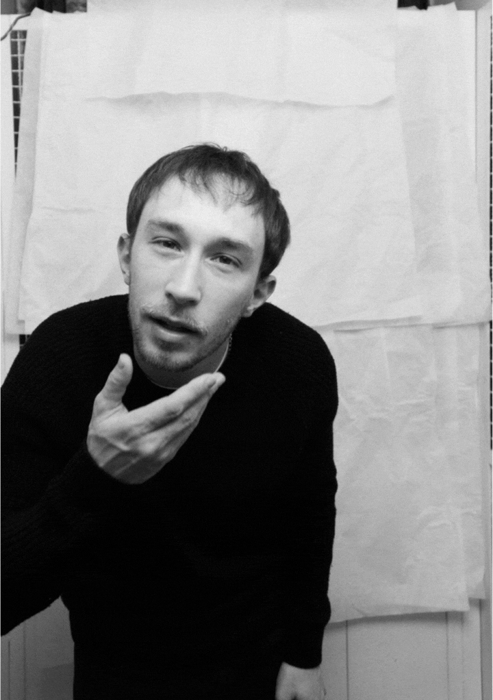 Black and white photo of a man with short hair and a beard, wearing a dark sweater, leaning forward with his hand near his face.