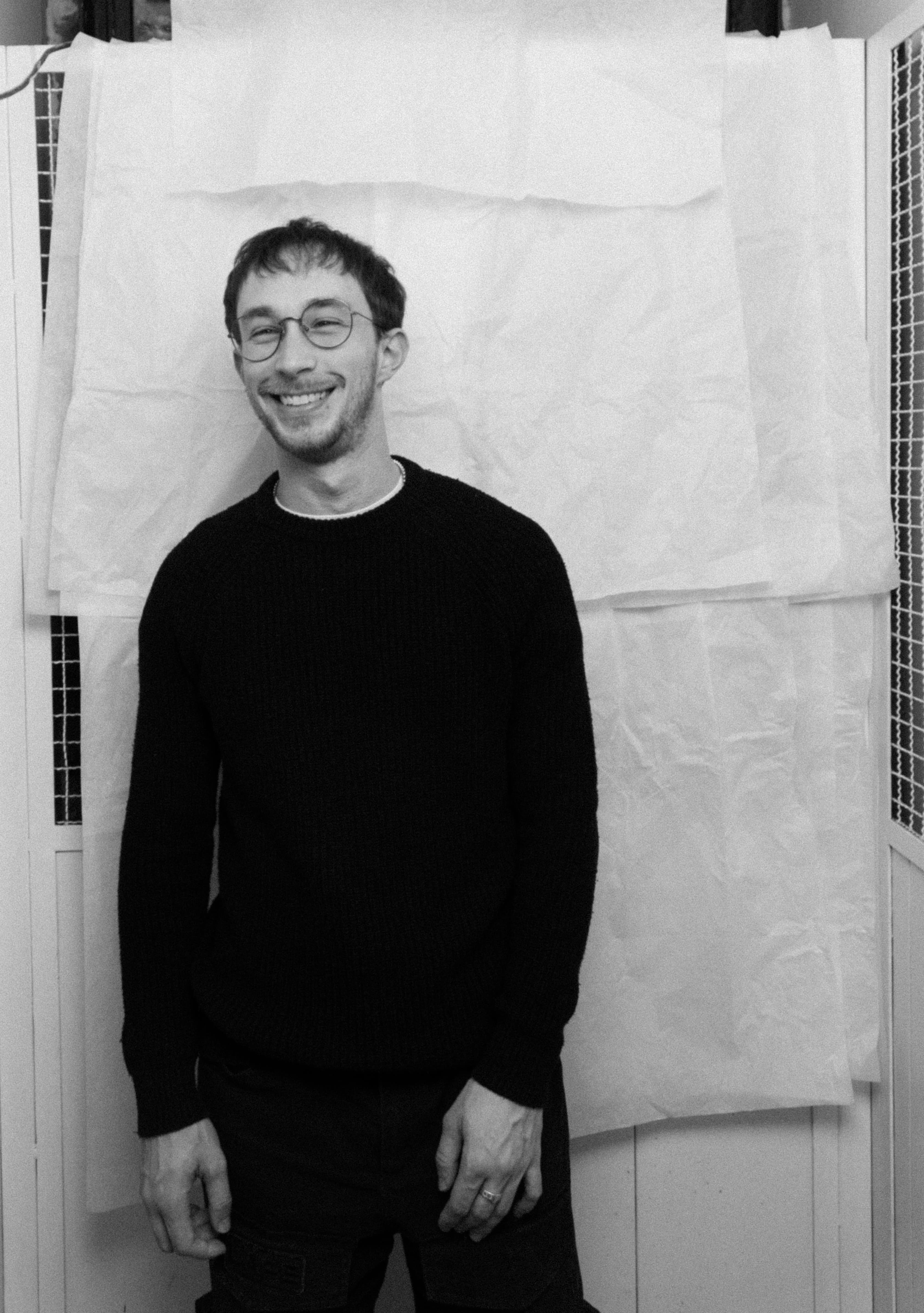 Smiling man wearing glasses and a black sweater standing against a wall with white fabric panels.