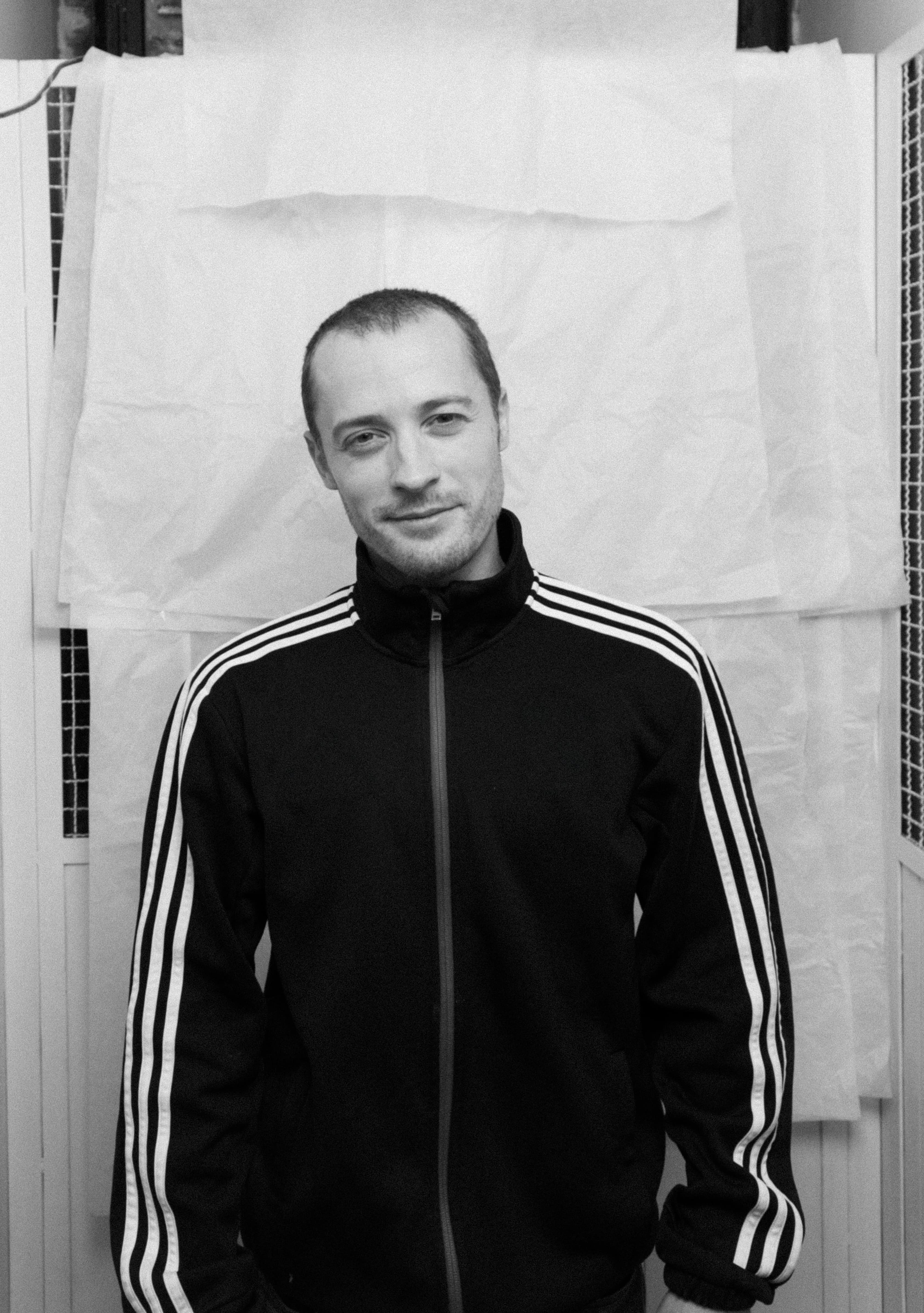 Portrait of a man wearing a black jacket with white stripes on the sleeves, standing in front of a white backdrop.
