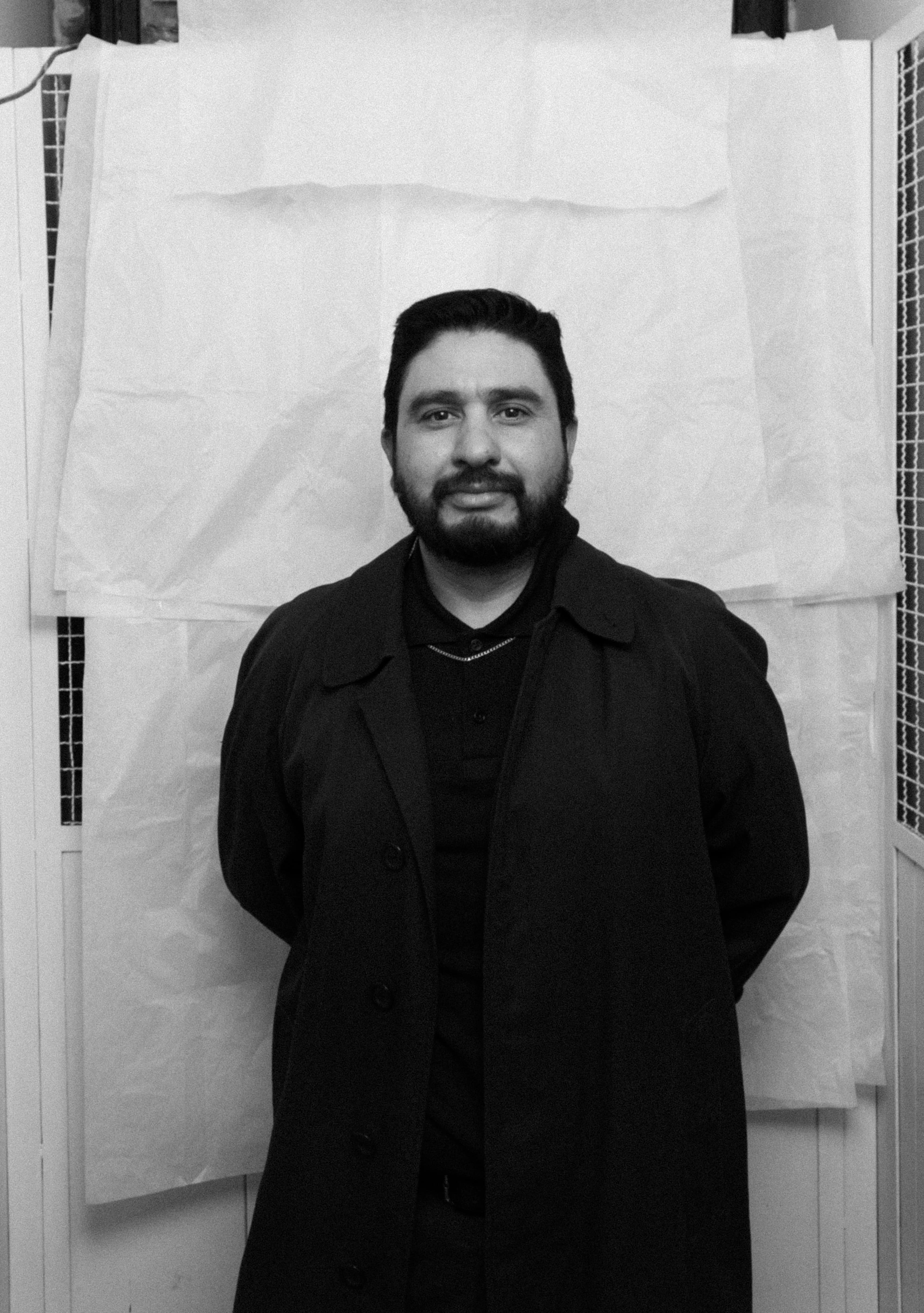 A bearded man wearing a dark coat and black shirt stands in front of a white textured background.