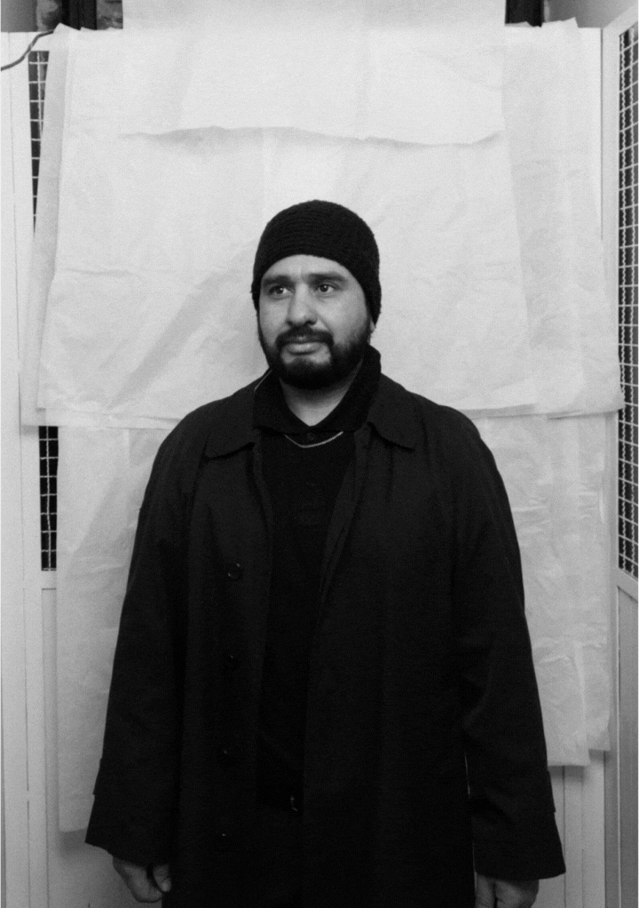Bearded man wearing a black coat and knit beanie standing against a white backdrop.