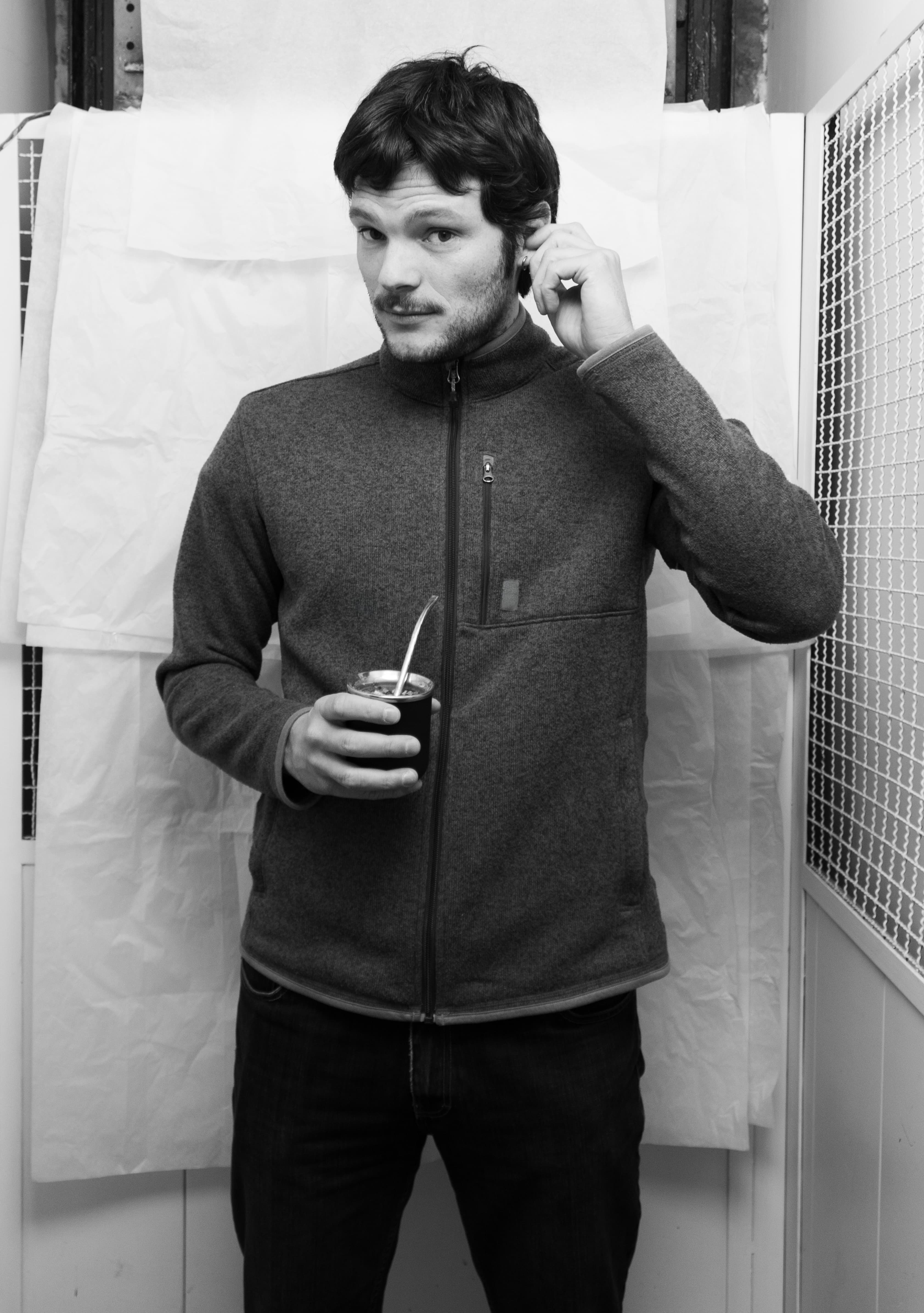 Man with short dark hair and a beard wearing a zip-up fleece jacket holding a mate cup with a straw while touching his ear.