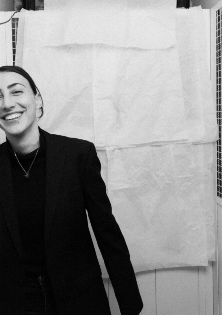Smiling person wearing a dark blazer and necklace, standing against a white textured background.