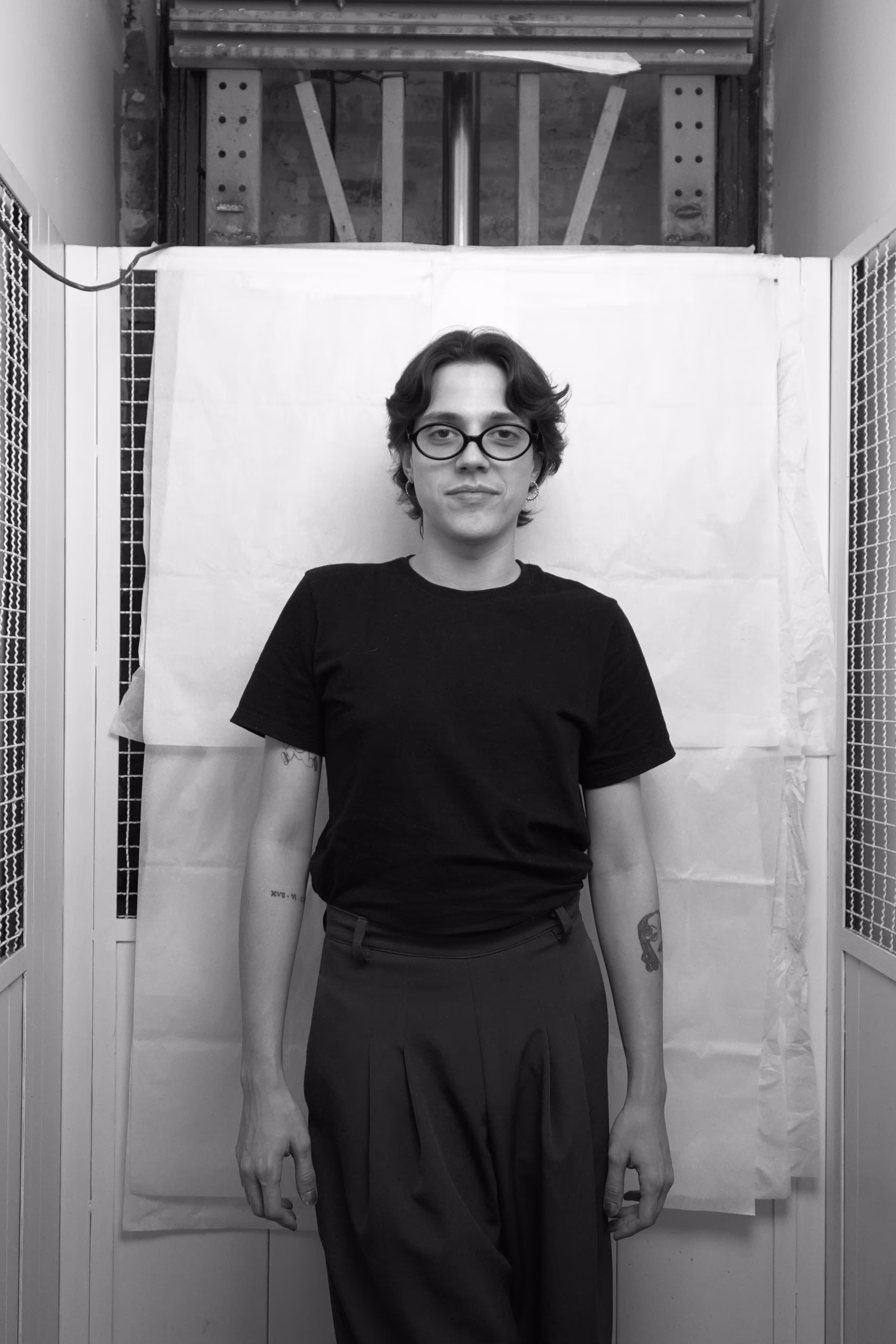 Portrait of a man with glasses and a beard standing with hands behind his back in front of a white textured backdrop.