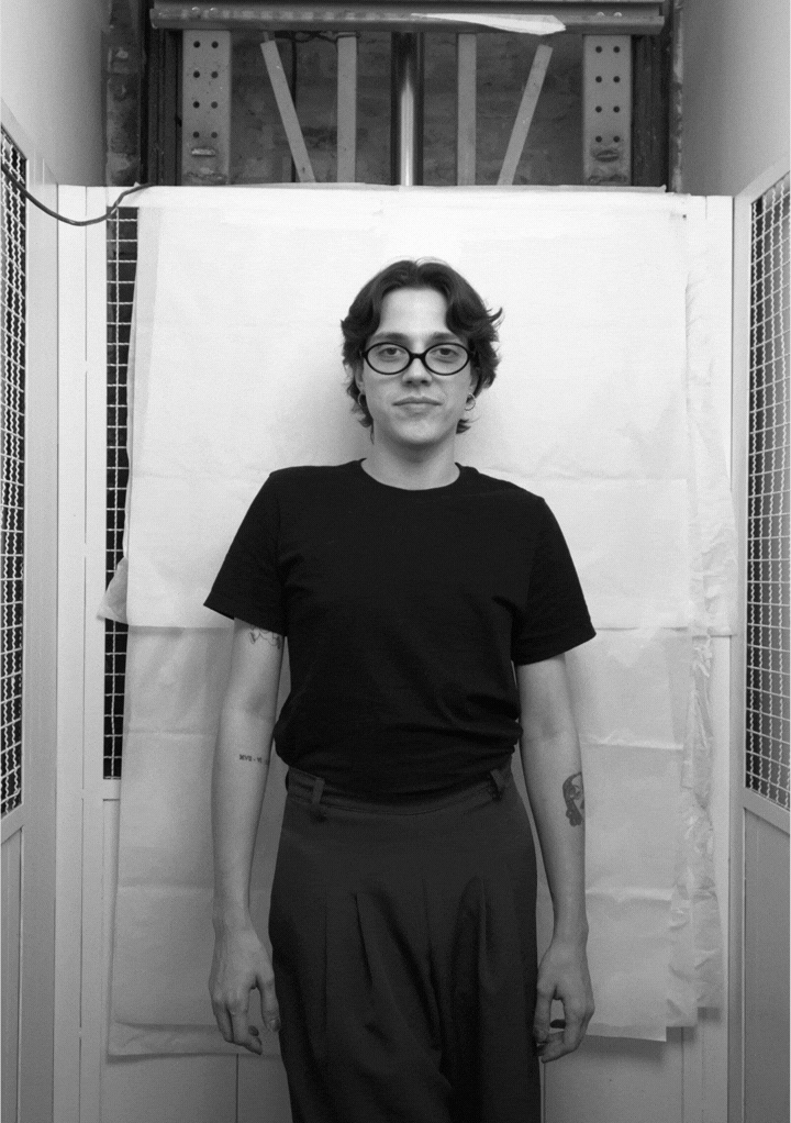 Black and white photo of a man with a beard holding glasses to his face, standing with crossed arms in front of a textured backdrop.