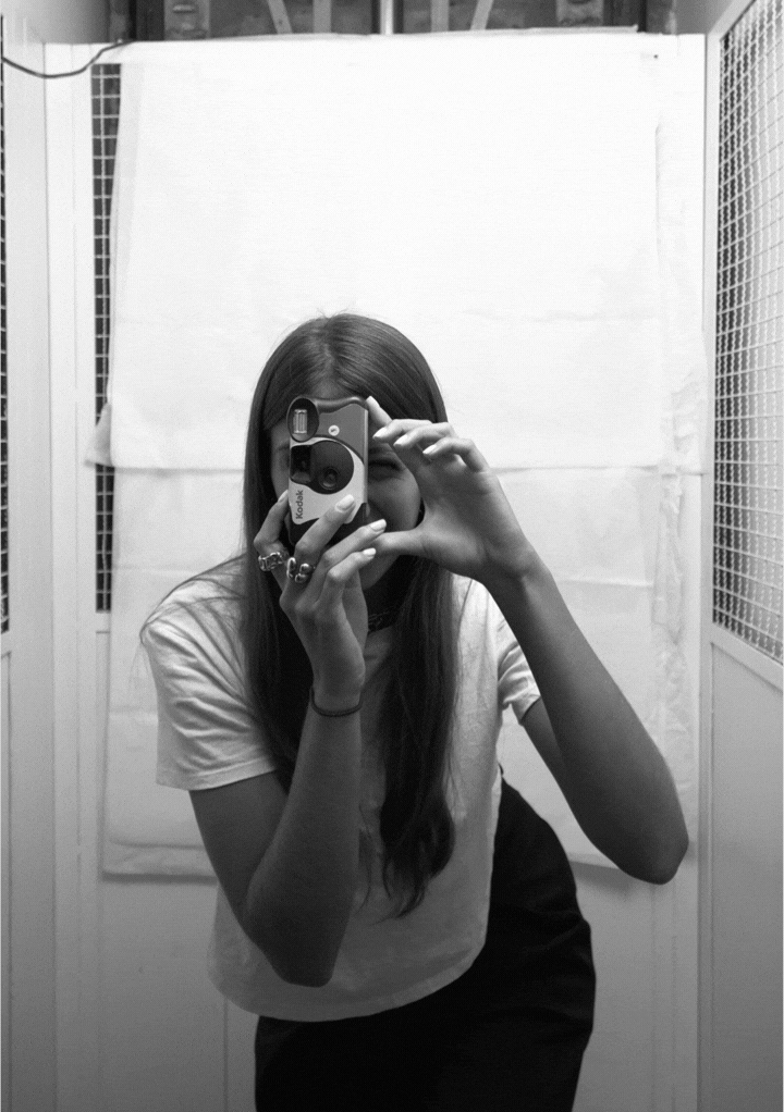 Black-and-white photo of a woman with shoulder-length hair wearing large white sunglasses, a choker, and a dark long-sleeve top, with a small toy animal placed on her head.