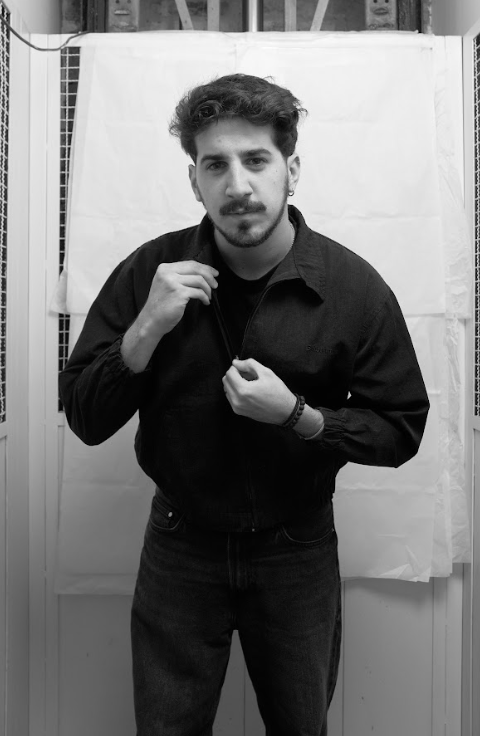 Smiling man with short dark hair and beard wearing a black t-shirt standing against a white paper backdrop in a black and white photo.