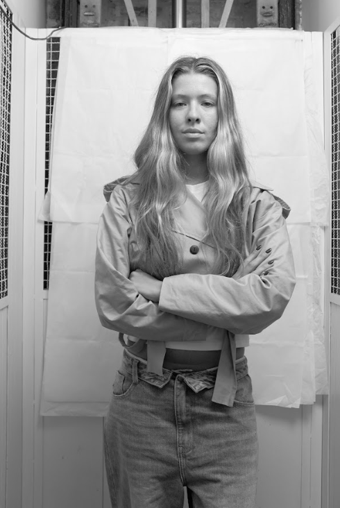 Black and white photo of a woman with curly hair wearing a black top and pants, standing with hands behind her back, looking upward thoughtfully.