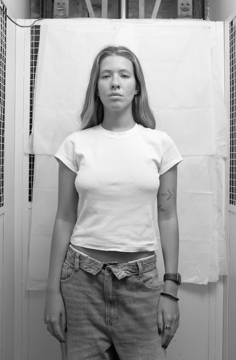 Black and white photo of a woman with curly hair wearing a black top and pants, standing with hands behind her back, looking upward thoughtfully.