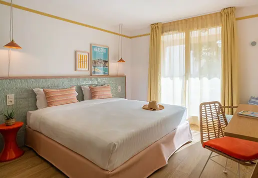 Chambre d'hôtel lumineuse avec un grand lit double, deux lampes suspendues, un fauteuil en rotin avec coussin orange, un bureau, et une grande fenêtre avec rideaux jaunes laissant entrer la lumière naturelle.