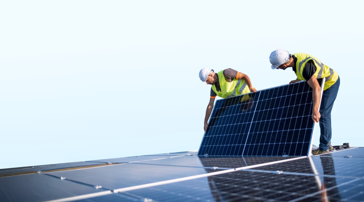 Two workers wearing safety helmets and reflective vests installing solar panels on a roof against a clear sky.
