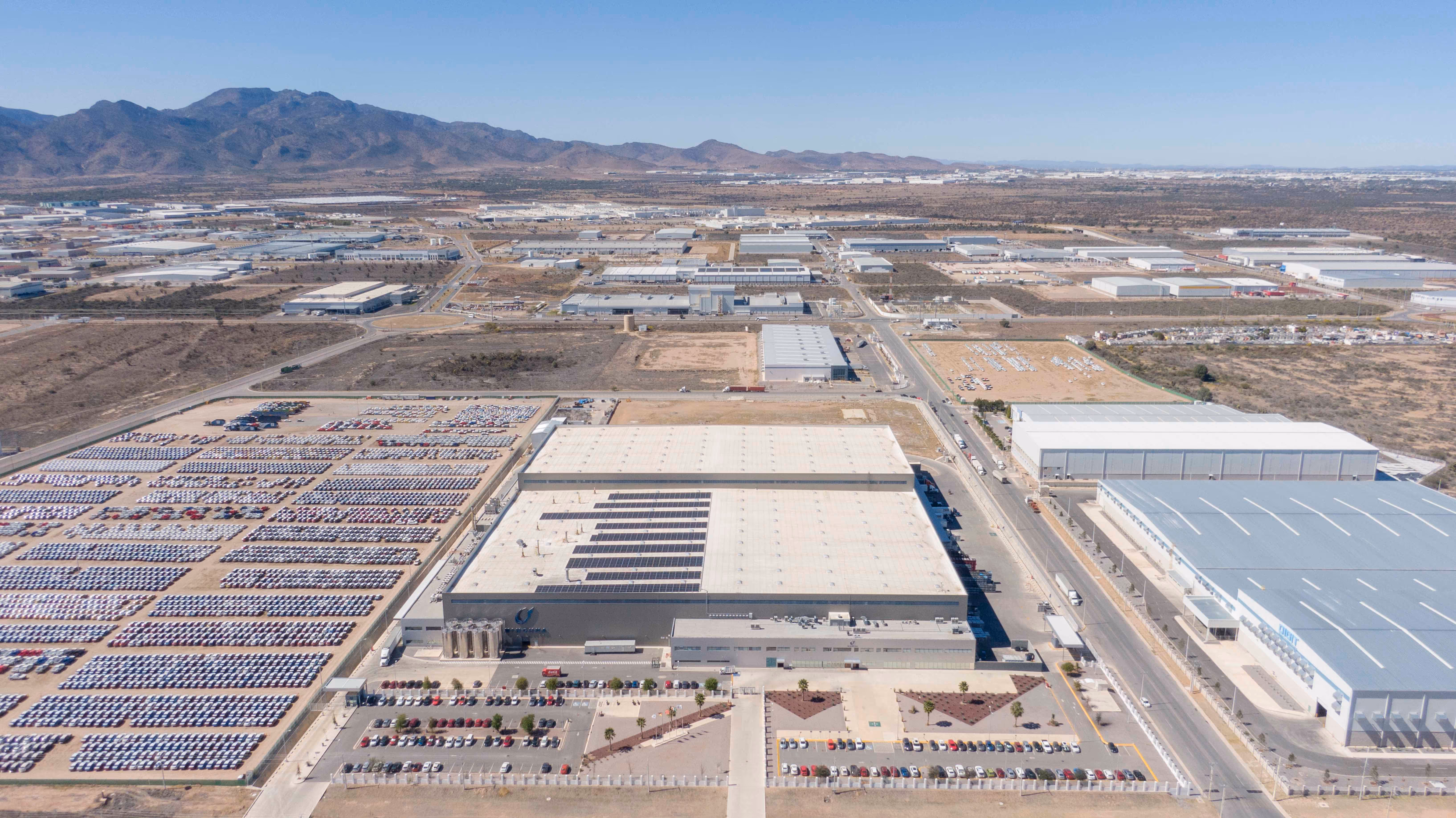 Aerial view of industrial buildings, large parking lots filled with cars, and distant mountains under clear sky.