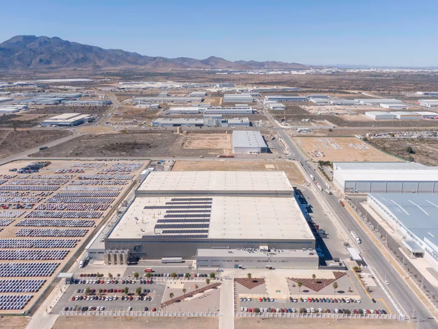 Aerial view of industrial buildings, large parking lots filled with cars, and distant mountains under clear sky.