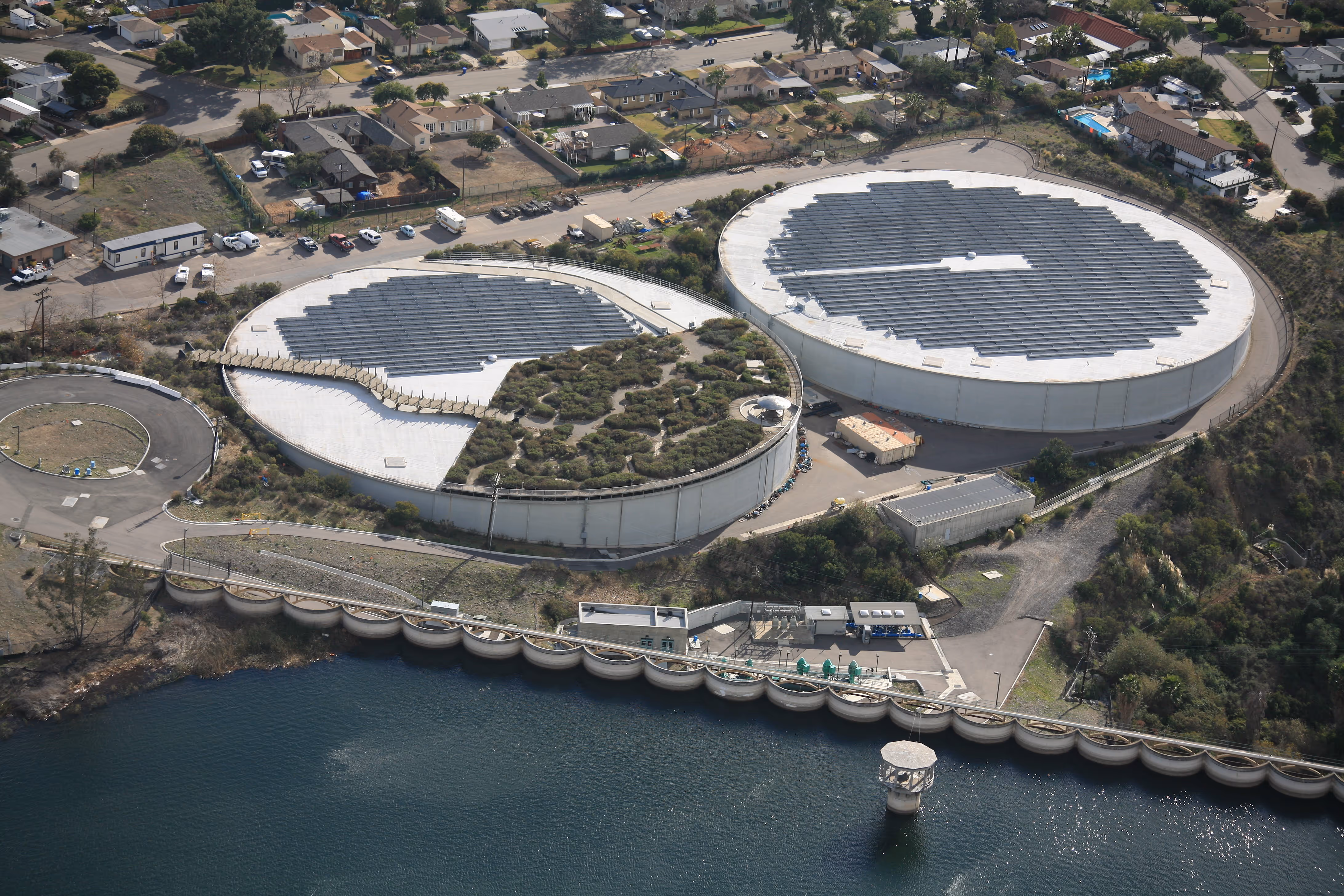 Aerial view of circular water tanks with solar panels and vegetation near a body of water and residential area.