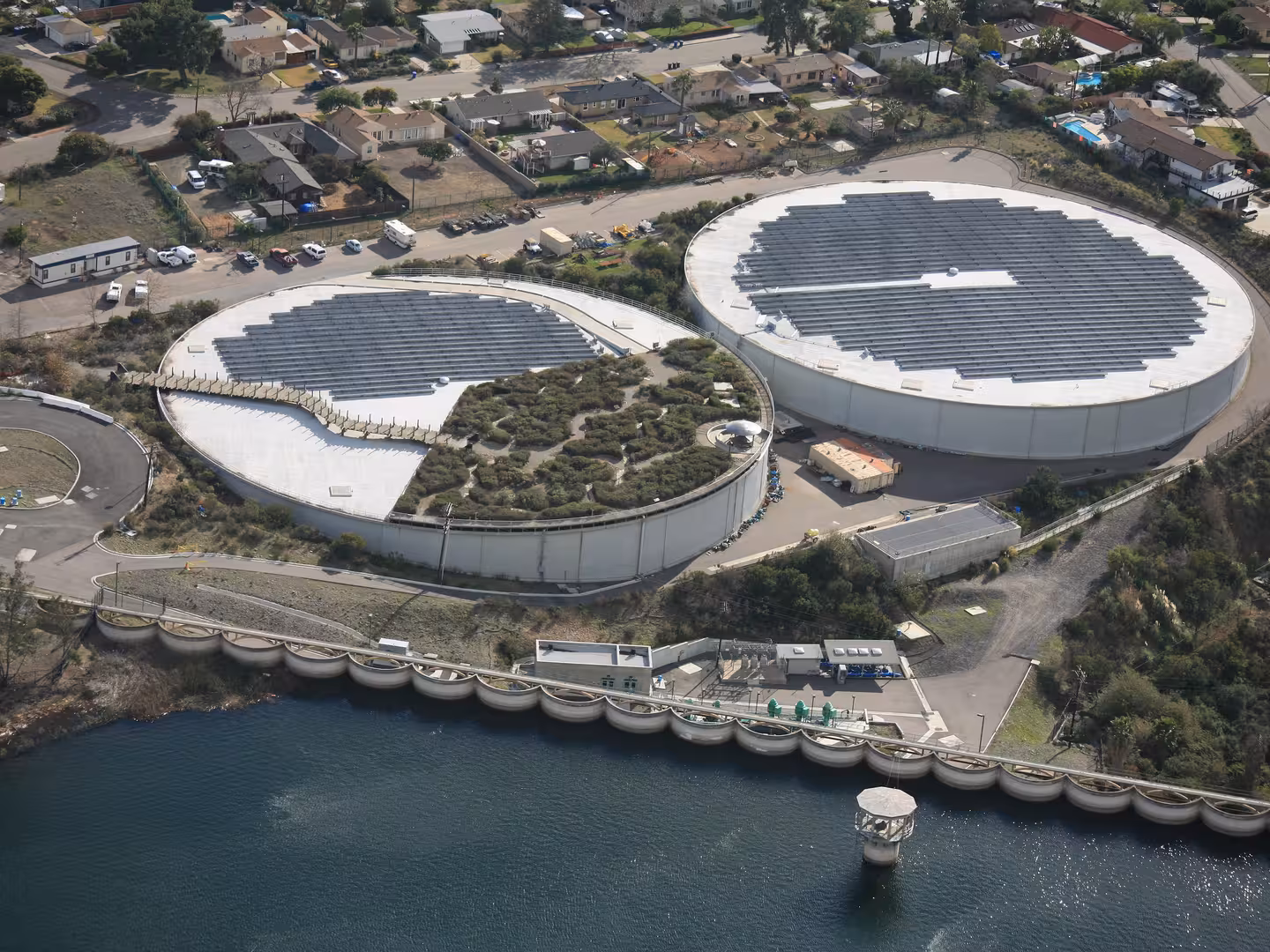 Aerial view of circular water tanks with solar panels and vegetation near a body of water and residential area.