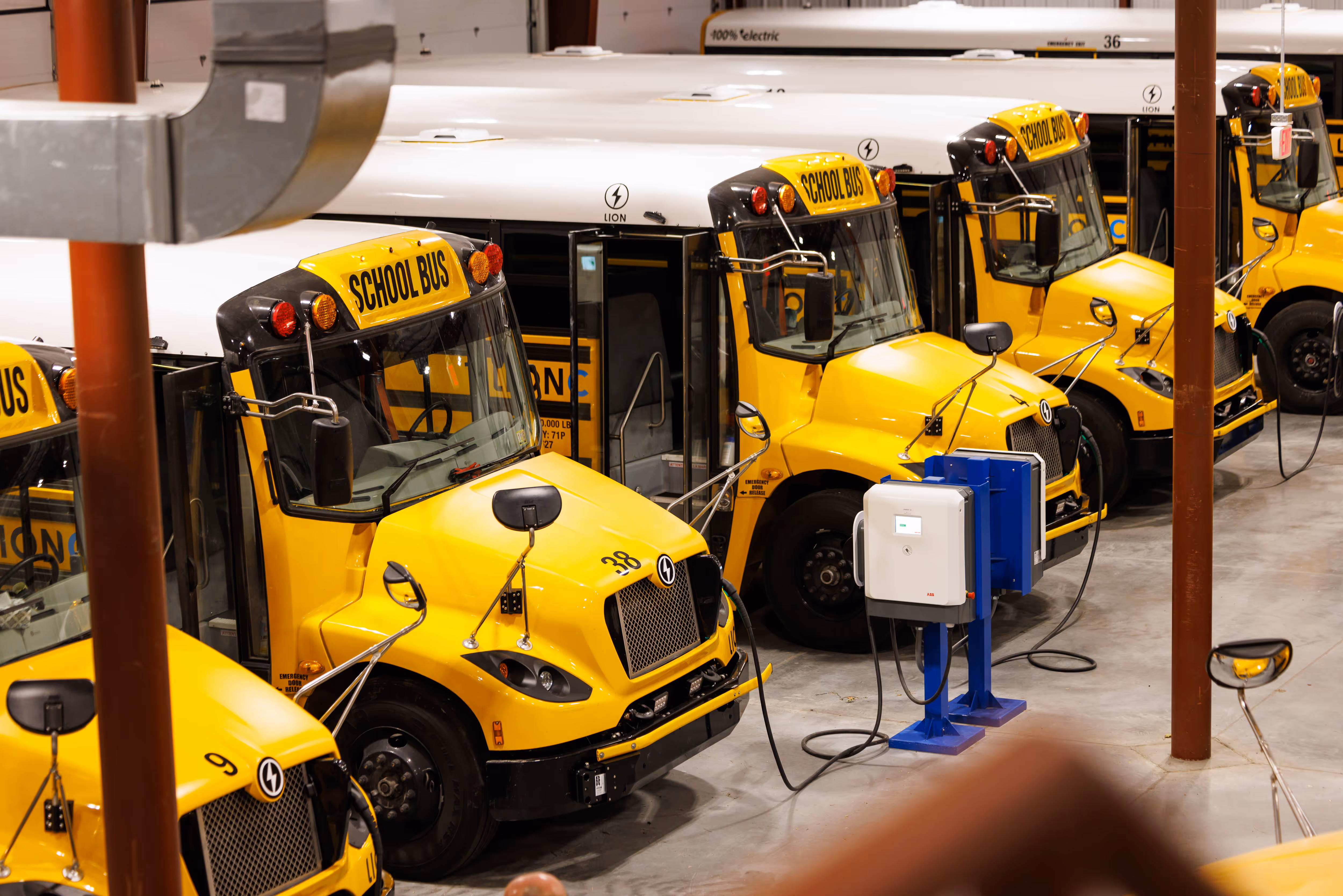 Yellow electric school buses parked indoors, plugged into blue charging stations.