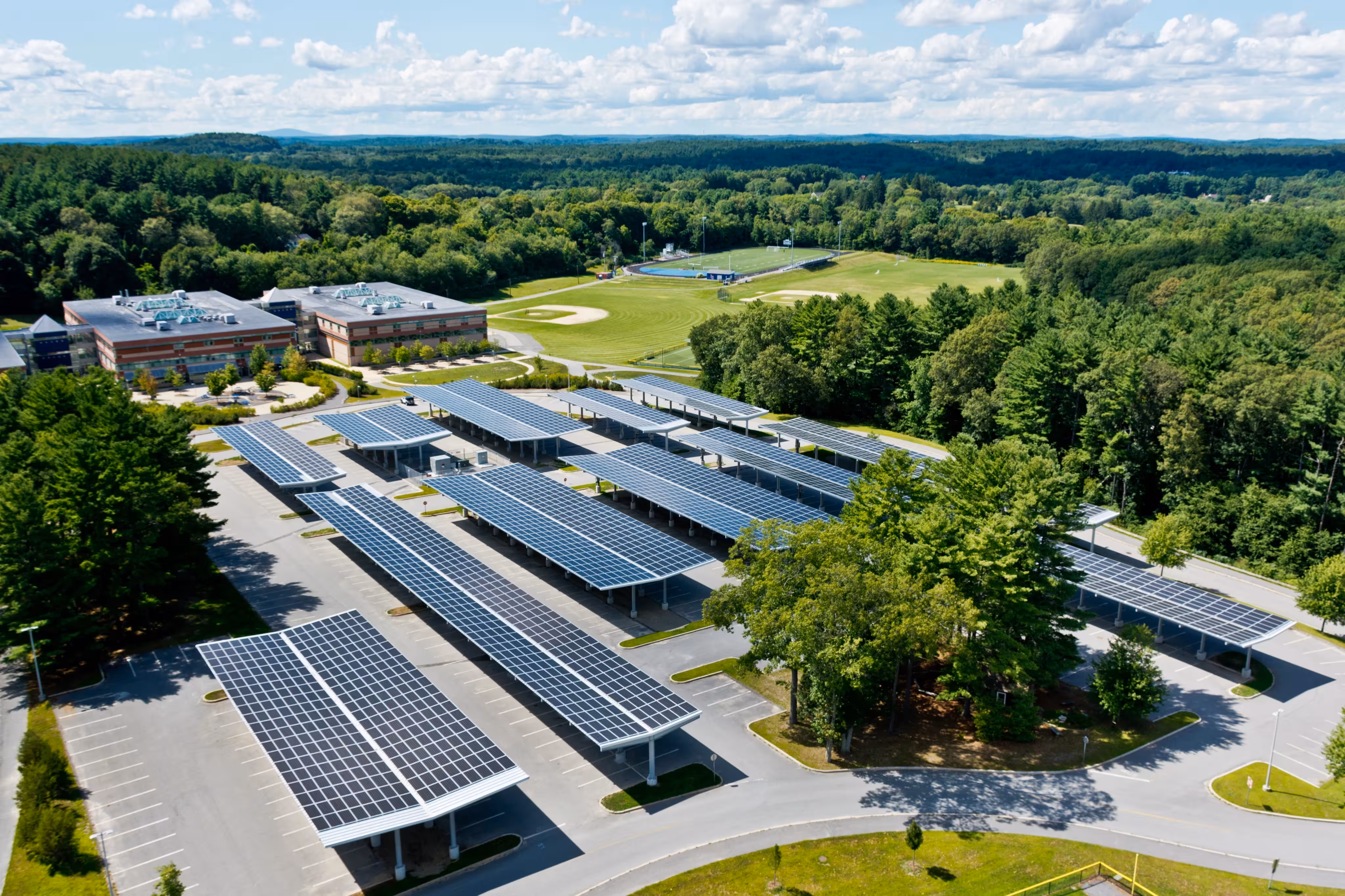 Aerial view of solar panel canopies covering a school parking lot with sports fields and forest in background.