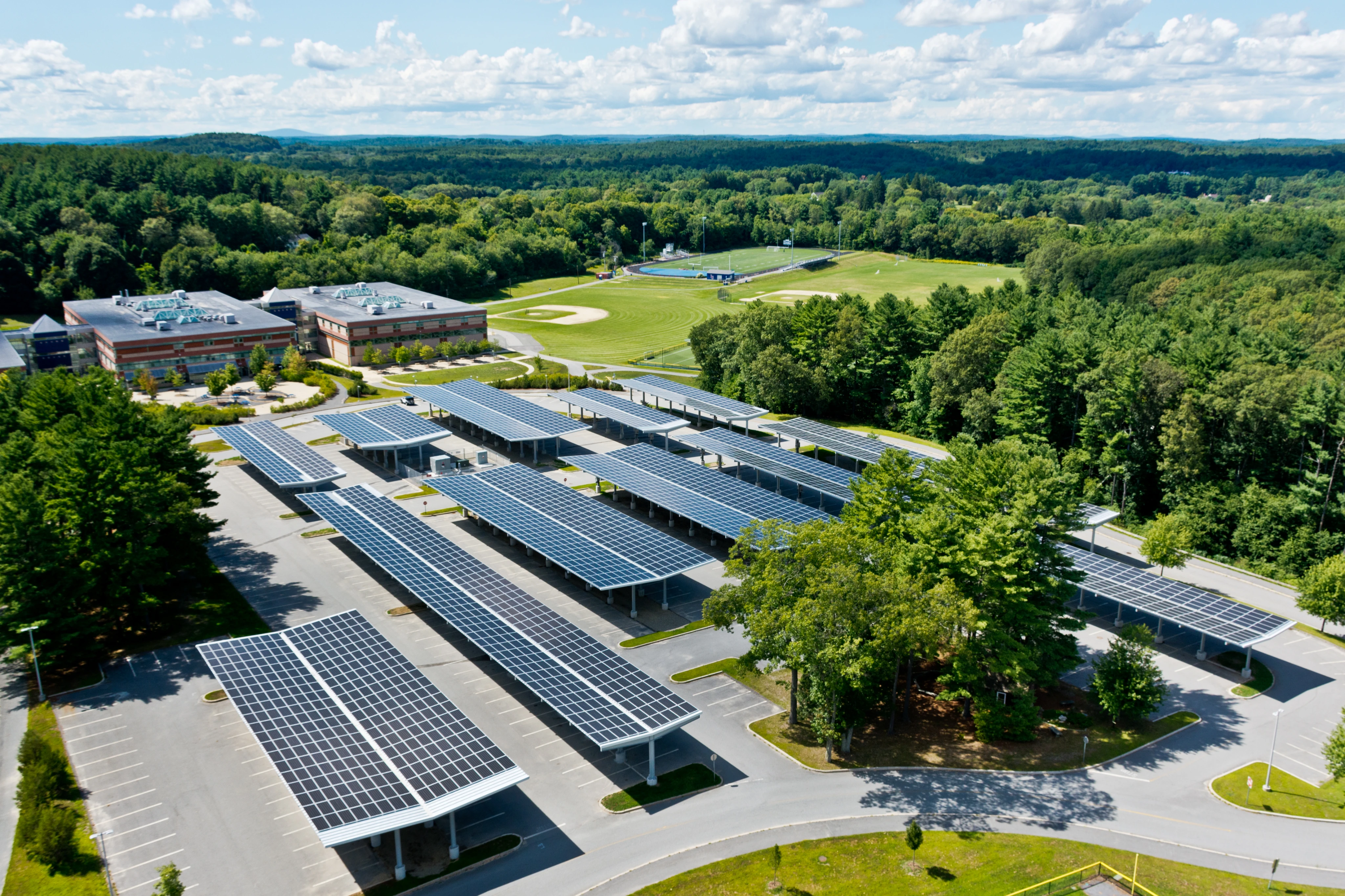 Aerial view of solar panel canopies covering a school parking lot with sports fields and forest in background.