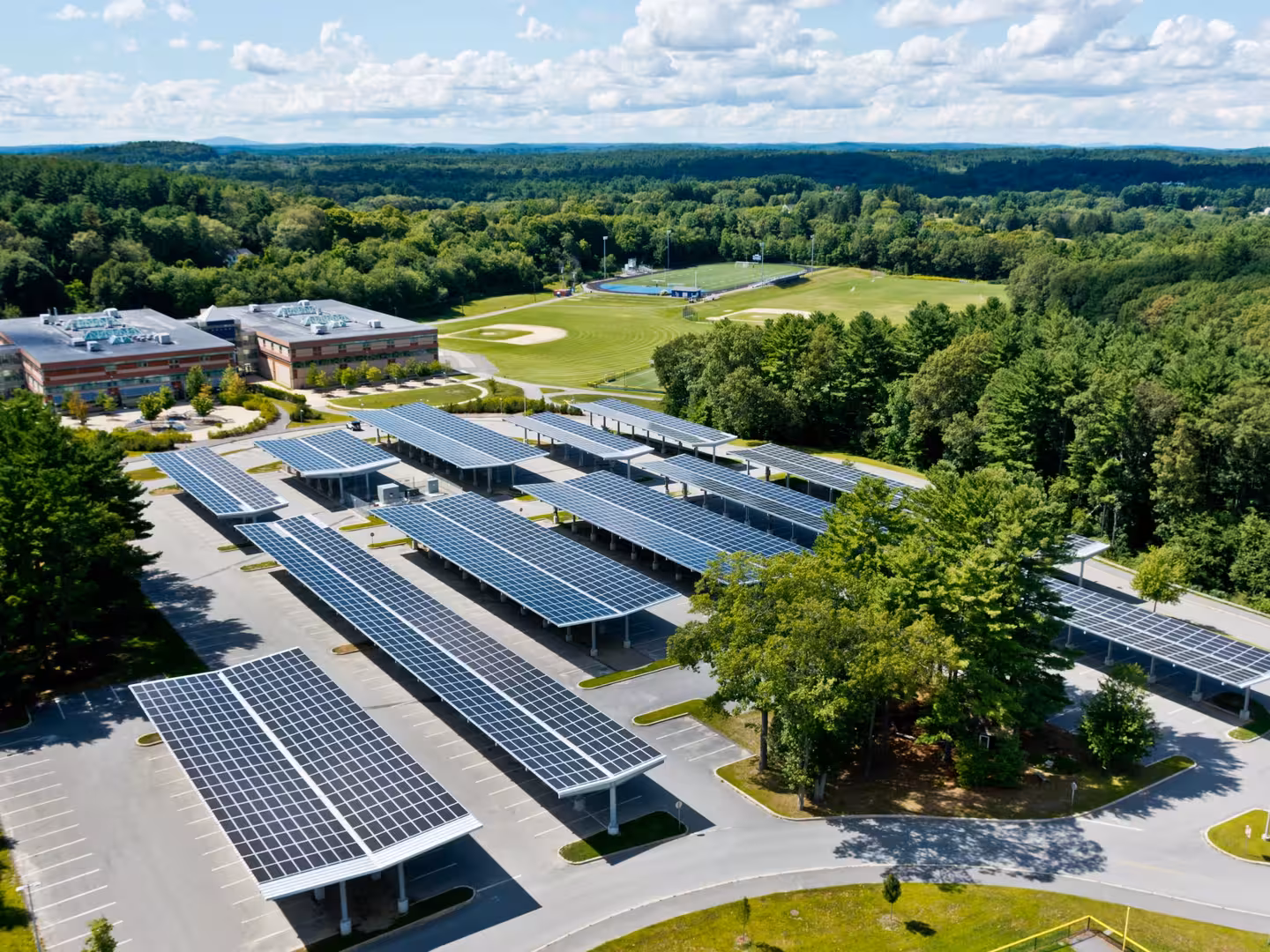 Aerial view of solar panel canopies covering a school parking lot with sports fields and forest in background.