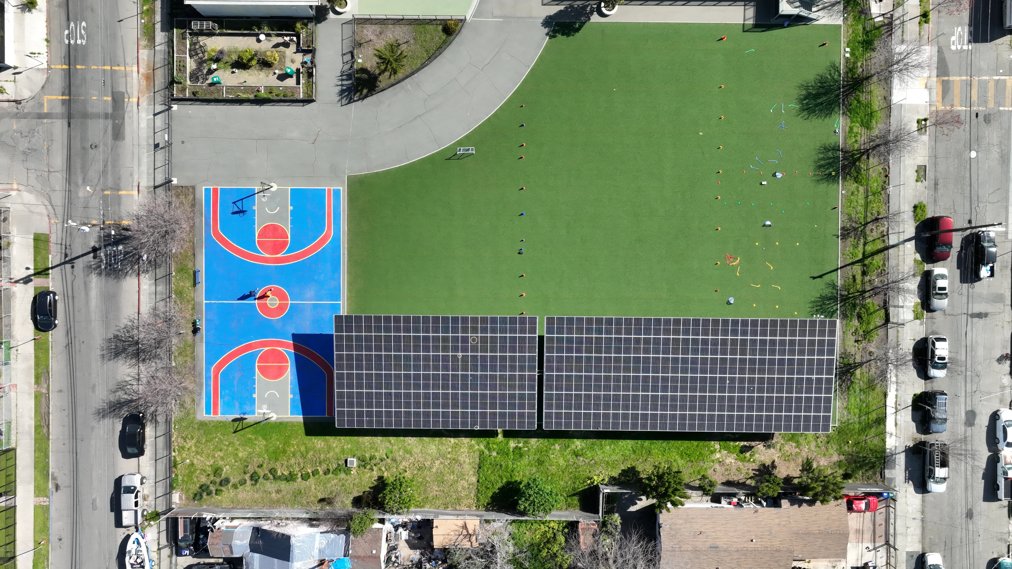 Aerial view of a blue and red basketball court next to a green field with cones and solar panels on a building roof.