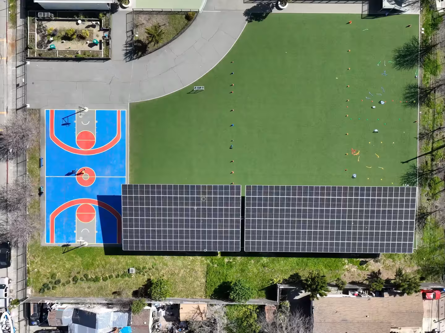 Aerial view of a blue and red basketball court next to a green field with cones and solar panels on a building roof.